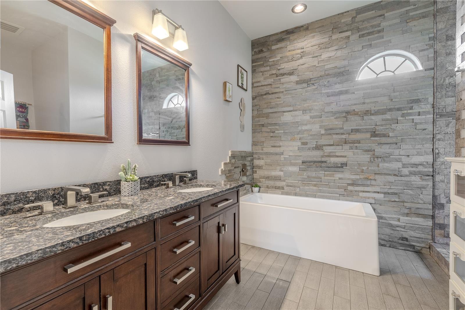 The primary ensuite bath features a mirrored dual sink wood vanity with granite countertop and down light fixtures. A modern soaking tub sits beside a stone accent wall.