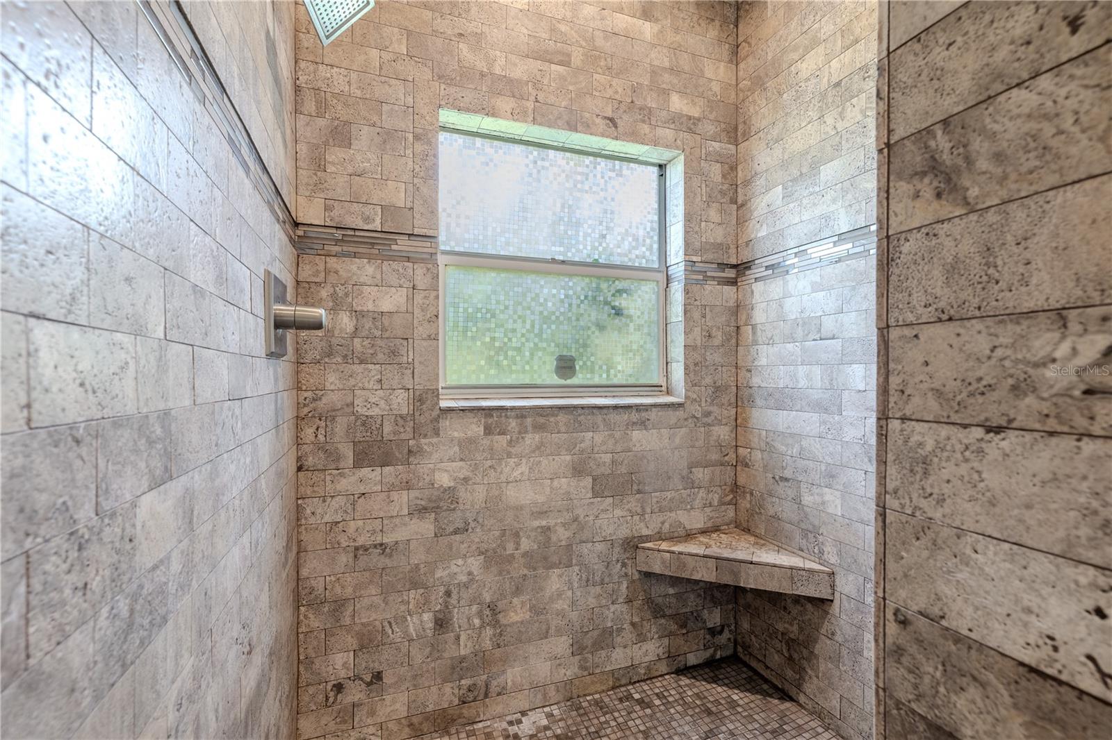 Indulge in the luxurious primary bath featuring a custom stone-tiled walk-in shower with dual shower heads for the ultimate spa experience. This thoughtfully designed shower includes a convenient built-in corner seat, decorative accent tile detailing, and a privacy window that bathes the space in natural light while ensuring complete privacy.