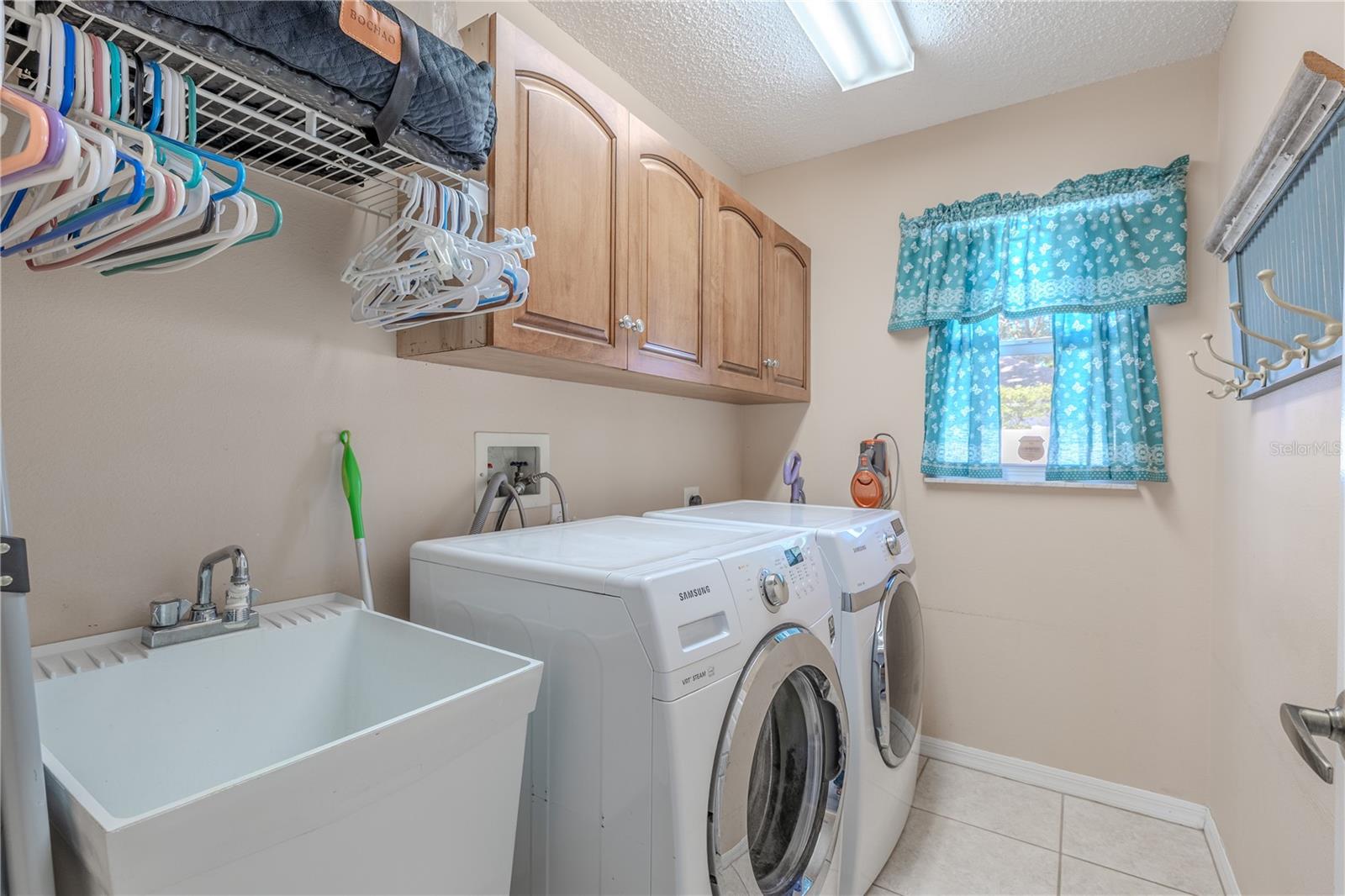 The laundry room features a large utility sink, perfect for cleaning larger items or hand-washing. Above, there are wooden cabinets providing ample storage space for laundry essentials and household supplies. A convenient wall-mounted shelf with a hanging rod offers additional organization options for clothes and accessories. The room is equipped with a washer and dryer, making laundry chores efficient and straightforward.
