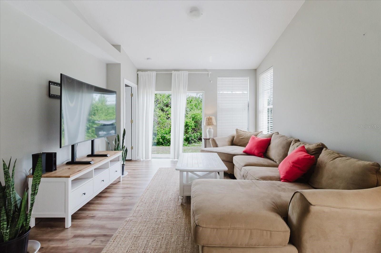 Open Concept Main Living Area With HIGH CEILINGS and Luxury Vinyl Floors, Sliding Glass Door Leads To Patio That Overlooks A Wood Area 2