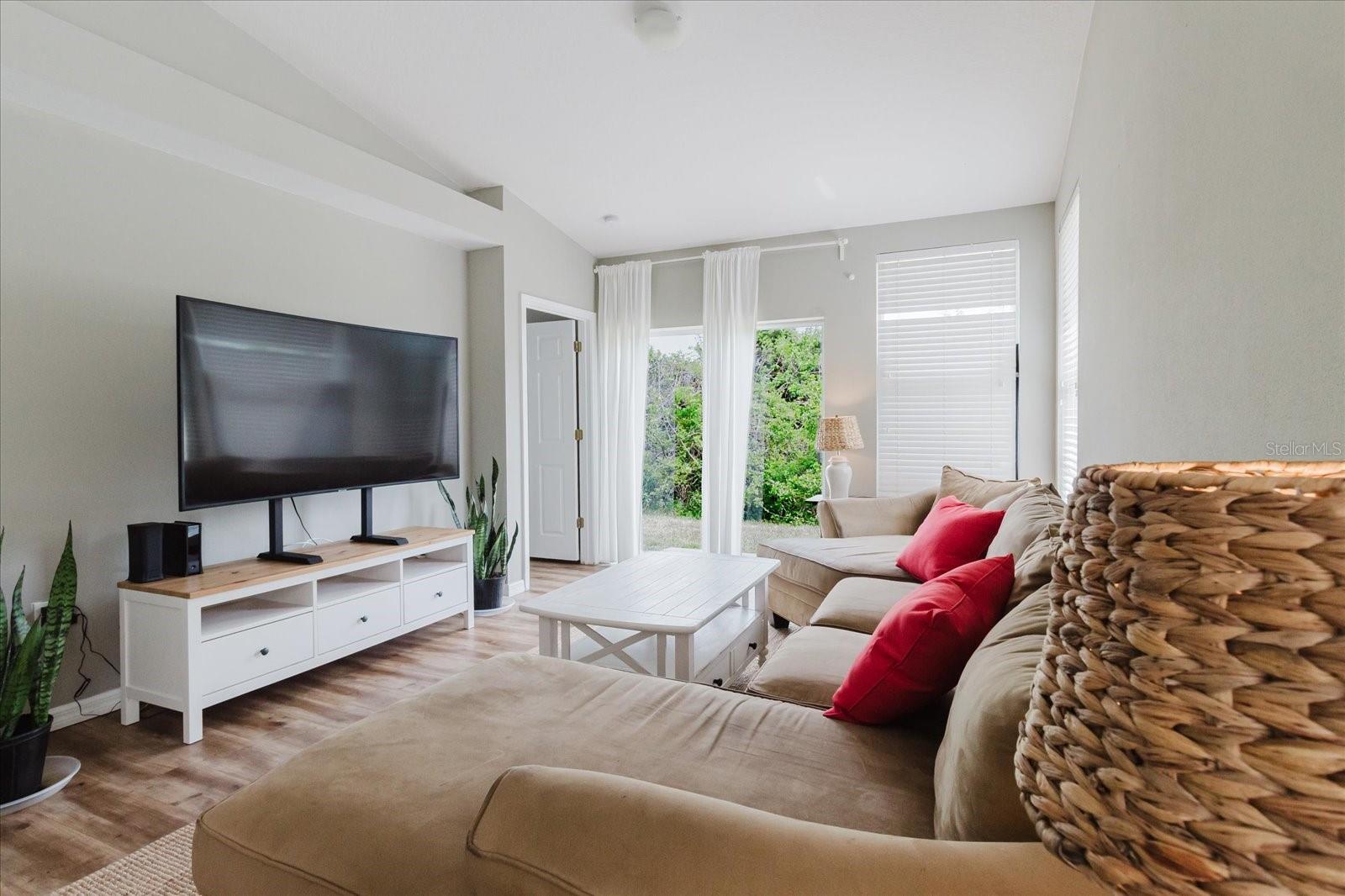 Open Concept Main Living Area With HIGH CEILINGS and Luxury Vinyl Floors, Sliding Glass Door Leads To Patio That Overlooks A Wood Area 3