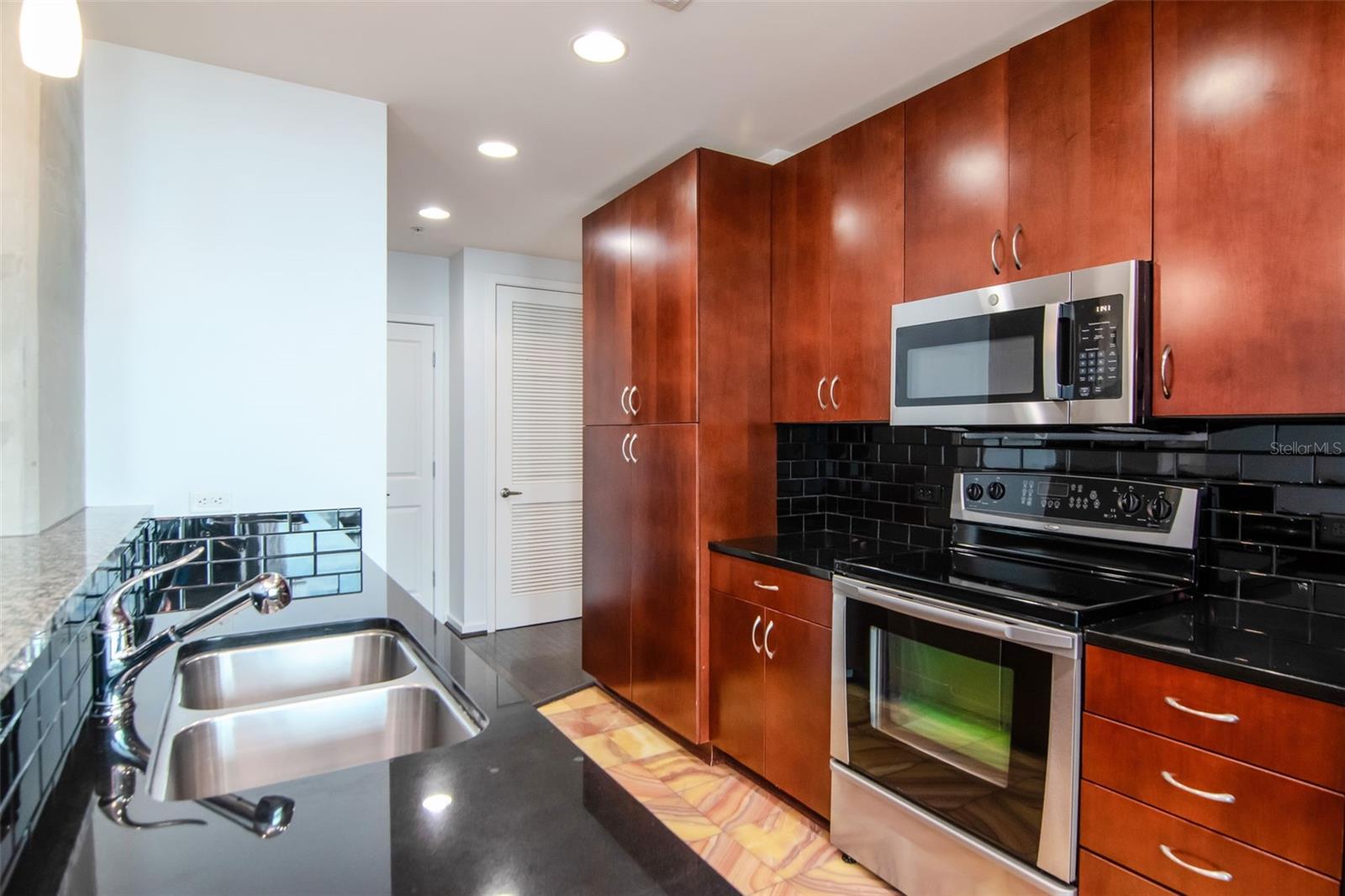 Sleek granite countertops, glossy black subway-tile backsplash, rich cherry-wood cabinetry, and stainless-steel appliances.