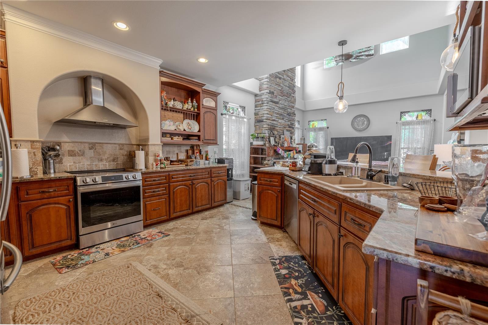 This well-appointed kitchen is the perfect blend of classic style and everyday functionality.Rich wood cabinetry with crown molding and glass-front accents provides generous storage, while granite countertops and a coordinating tile backsplash add timeless elegance. A custom arched range hood creates a stunning focal point above the stainless steel oven, complemented by matching appliances including a French door refrigerator and dishwasher. The open layout flows effortlessly into the living area, making it easy to entertain while cooking. Additional features include a deep dual-basin sink, built-in microwave niche, under-cabinet lighting, pendant accents, and abundant prep space for the home chef.