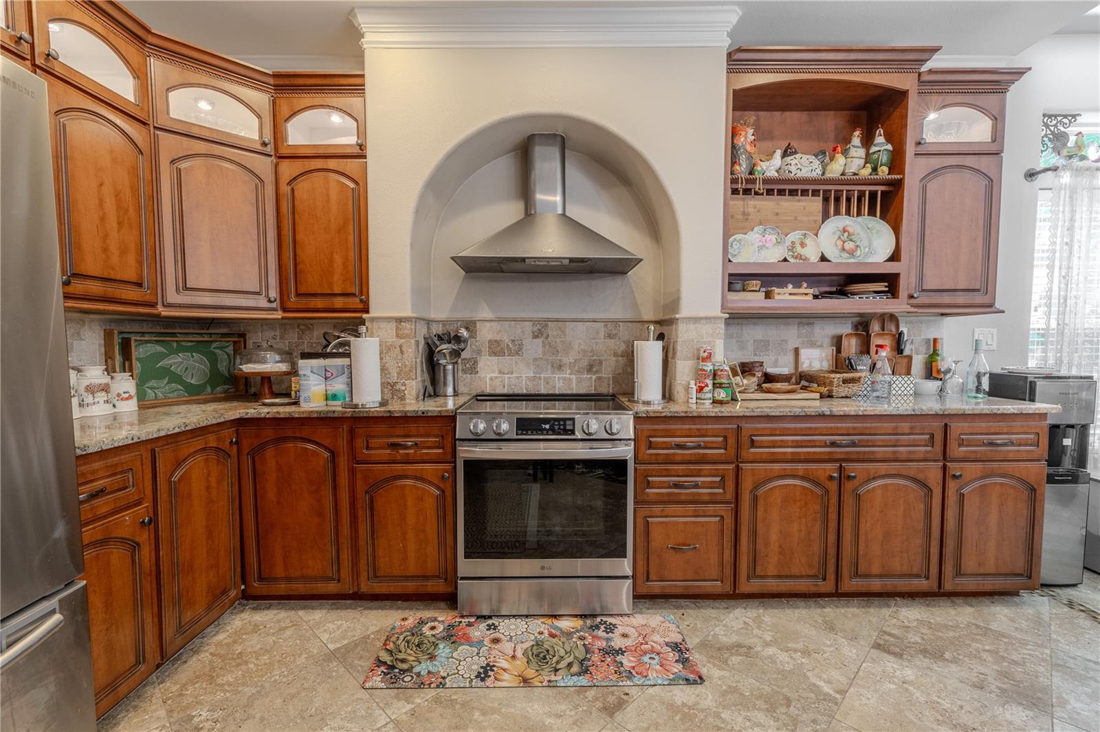 This well-appointed kitchen is the perfect blend of classic style and everyday functionality.Rich wood cabinetry with crown molding and glass-front accents provides generous storage, while granite countertops and a coordinating tile backsplash add timeless elegance. A custom arched range hood creates a stunning focal point above the stainless steel oven, complemented by matching appliances including a French door refrigerator and dishwasher. The open layout flows effortlessly into the living area, making it easy to entertain while cooking. Additional features include a deep dual-basin sink, built-in microwave niche, under-cabinet lighting, pendant accents, and abundant prep space for the home chef.