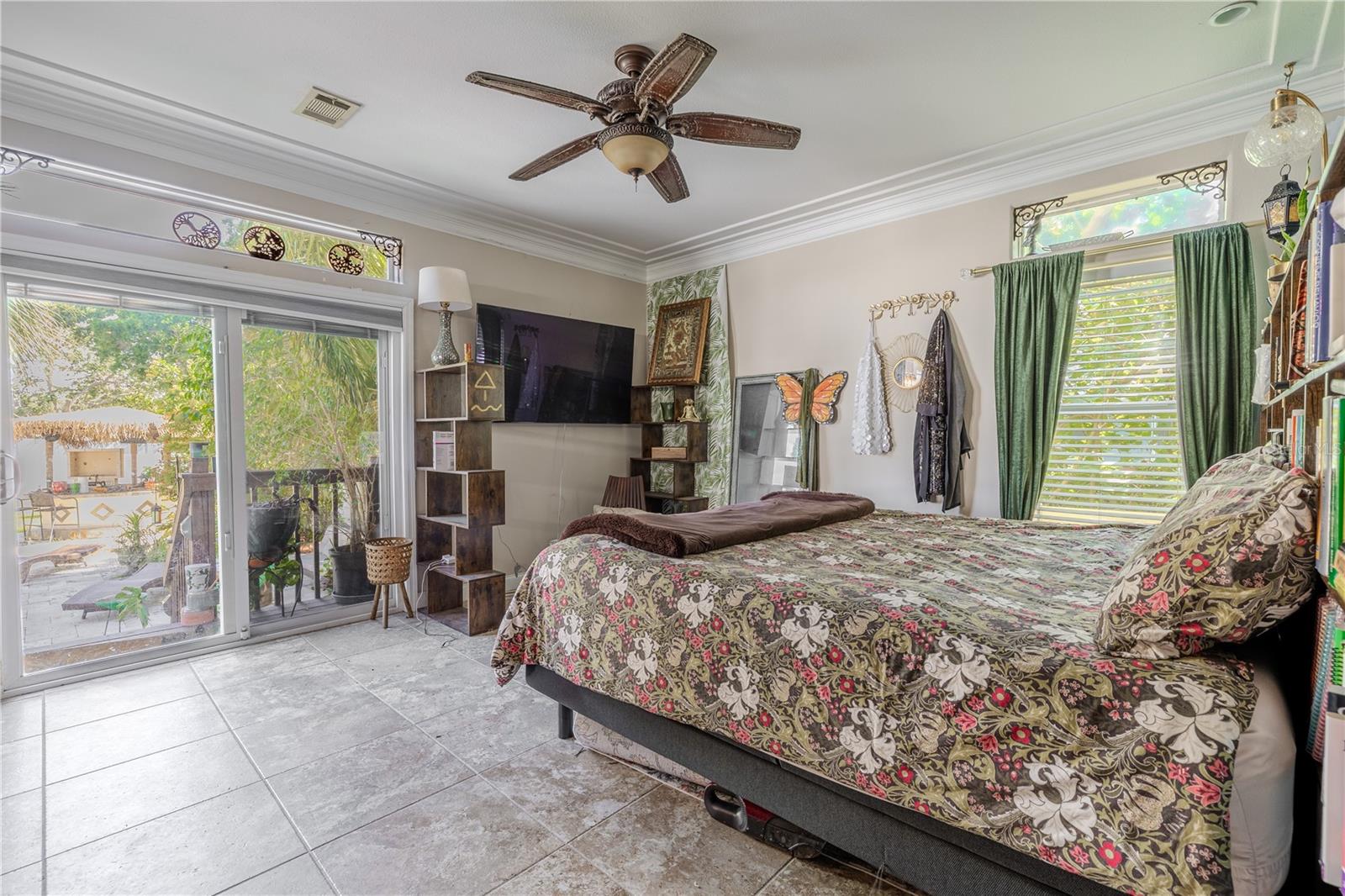 he spacious first-floor primary suite offers a peaceful retreat with direct access to the backyard oasis.?This light-filled room features crown molding, a designer ceiling fan, and large windows topped with decorative transoms that invite in natural light while maintaining privacy. Sliding glass doors lead directly to the patio, creating a seamless indoor-outdoor connection. The suite includes a generous walk-in closet and a private ensuite bath, offering comfort, convenience, and privacy—all just steps from the heart of the home.Primary bedroom.