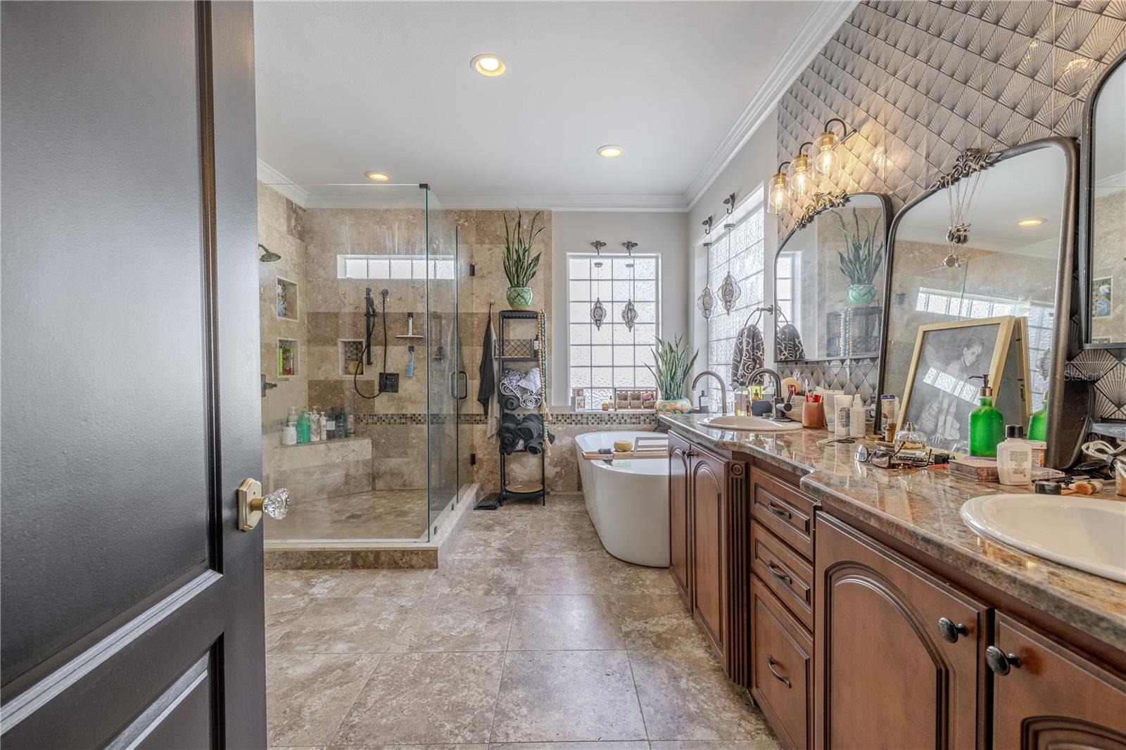 The luxurious primary ensuite bath offers a spa-like retreat with thoughtful design and upscale finishes throughout.Unwind in the freestanding soaking tub framed by two large glass block windows that fill the space with soft natural light while maintaining privacy. The oversized walk-in shower features floor-to-ceiling tile, a built-in bench, dual shower heads, and recessed niches for storage. A dual-sink vanity with rich wood cabinetry and granite countertops offers ample space for morning routines, highlighted by designer lighting and a textured accent wall with three arched mirrors. With its serene color palette, high-end fixtures, and stylish touches—including a tropical accent wall—this ensuite is the perfect blend of function and indulgence.