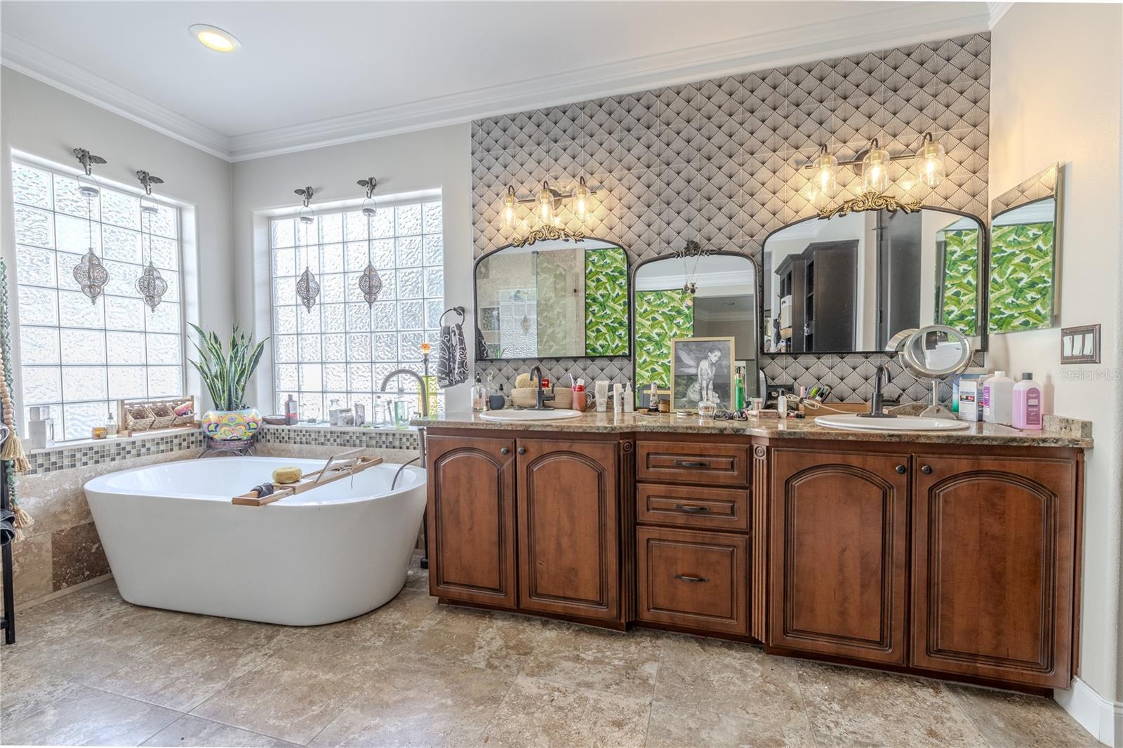Primary bathroomThe luxurious primary ensuite bath offers a spa-like retreat with thoughtful design and upscale finishes throughout.Unwind in the freestanding soaking tub framed by two large glass block windows that fill the space with soft natural light while maintaining privacy. The oversized walk-in shower features floor-to-ceiling tile, a built-in bench, dual shower heads, and recessed niches for storage. A dual-sink vanity with rich wood cabinetry and granite countertops offers ample space for morning routines, highlighted by designer lighting and a textured accent wall with three arched mirrors. With its serene color palette, high-end fixtures, and stylish touches—including a tropical accent wall—this ensuite is the perfect blend of function and indulgence.