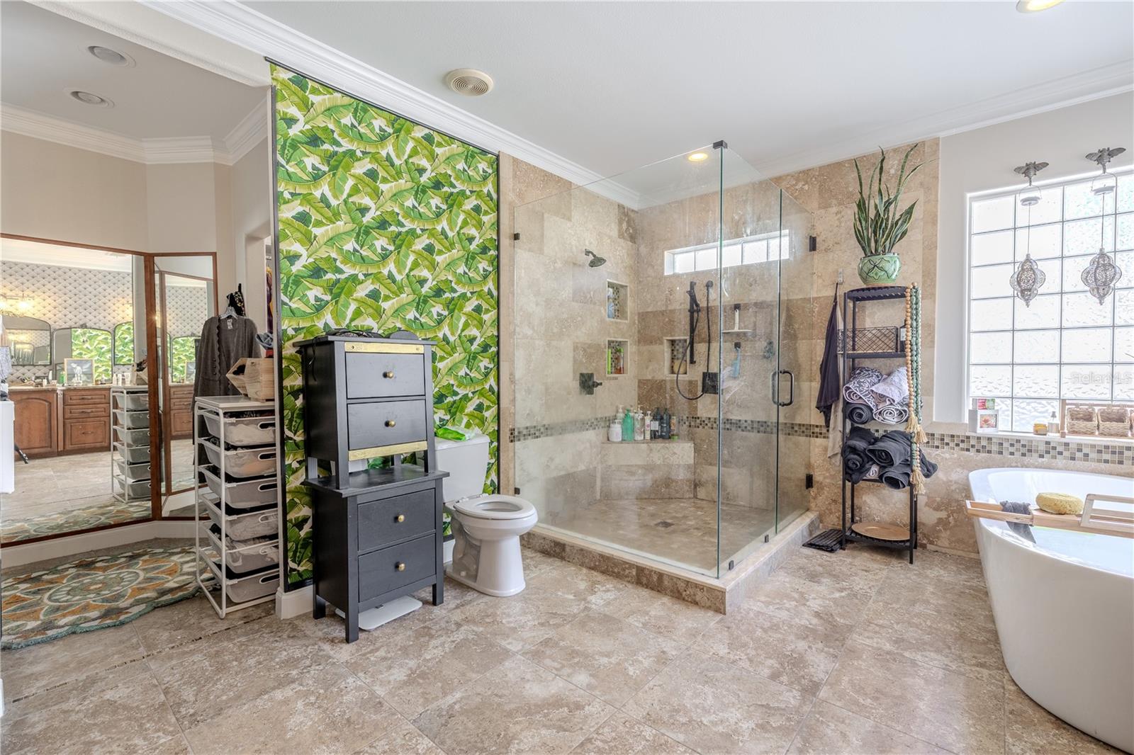 The luxurious primary ensuite bath offers a spa-like retreat with thoughtful design and upscale finishes throughout.Unwind in the freestanding soaking tub framed by two large glass block windows that fill the space with soft natural light while maintaining privacy. The oversized walk-in shower features floor-to-ceiling tile, a built-in bench, dual shower heads, and recessed niches for storage. A dual-sink vanity with rich wood cabinetry and granite countertops offers ample space for morning routines, highlighted by designer lighting and a textured accent wall with three arched mirrors. With its serene color palette, high-end fixtures, and stylish touches—including a tropical accent wall—this ensuite is the perfect blend of function and indulgence.
