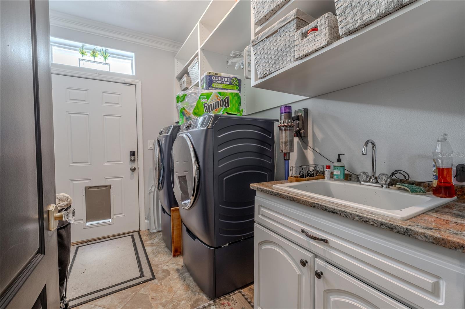 The well-designed laundry room offers both style and function with granite countertops, a deep utility sink, and upper open shelving for easy organization.Equipped with a sleek front-loading washer and dryer set on storage pedestals, this space makes laundry day a breeze. A door with built-in pet access leads to the exterior, while a transom window allows natural light to brighten the room. Whether you're managing daily chores or tackling weekend projects, this laundry area is both practical and thoughtfully appointed.