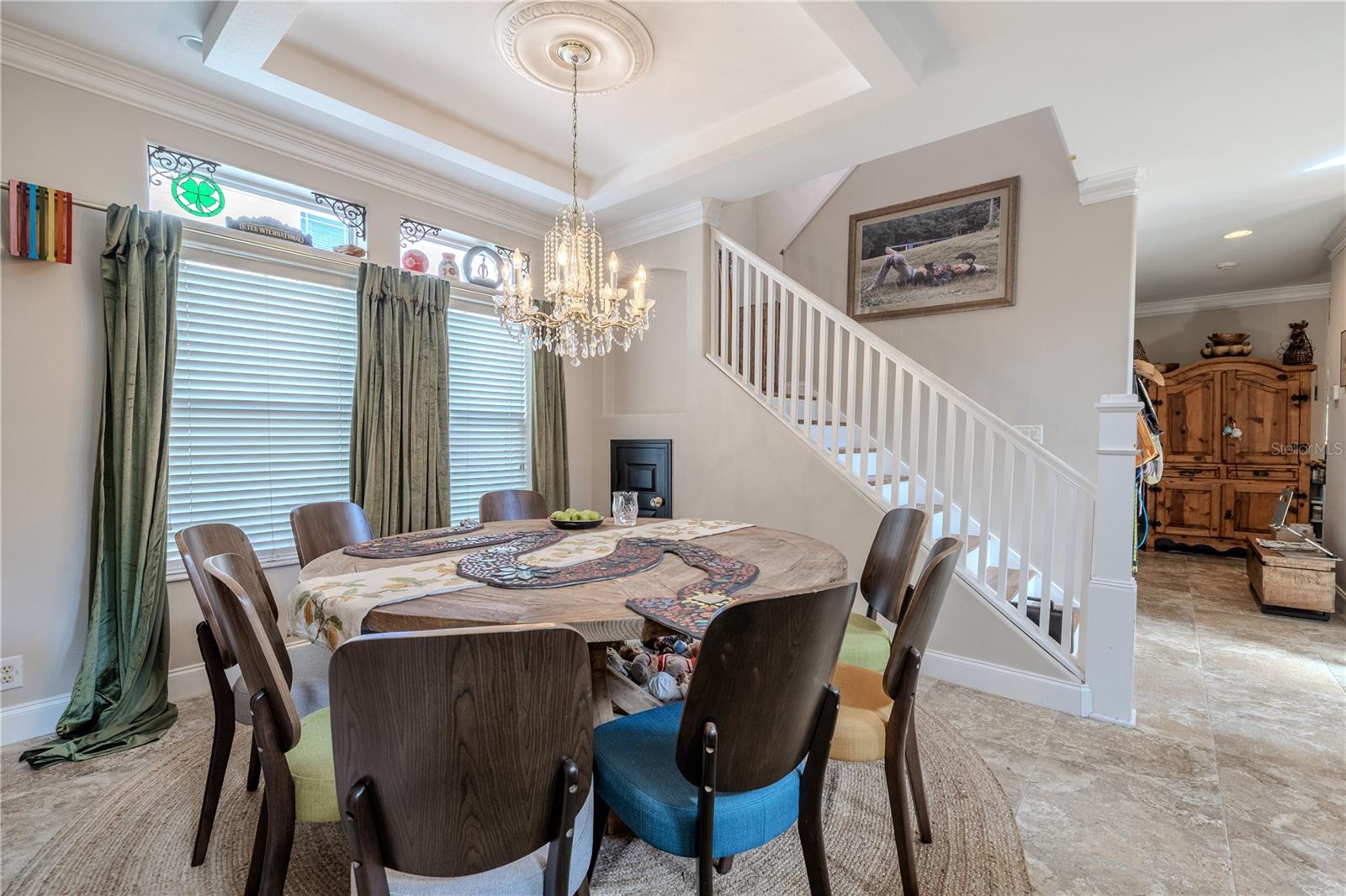 The elegant dining room is a true centerpiece of the home, featuring a statement crystal chandelier, classic crown molding, and a tray ceiling with a decorative medallion. Natural light pours in through tall windows topped with charming transom accents and wrought iron scrollwork, while floor-length drapes add a touch of drama. The space comfortably accommodates a large table, making it ideal for family gatherings or entertaining. Located just off the main entry and open to the flow of the home, this dining area blends style and function effortlessly.
