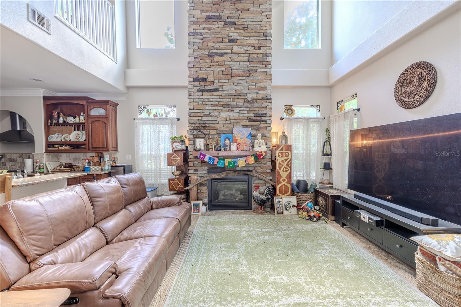 A dramatic two-story living room that blends comfort with architectural flair.Soaring ceilings and clerestory windows flood the space with natural light, while a stunning floor-to-ceiling stacked stone gas fireplace creates a cozy and commanding focal point. Custom transom windows with decorative iron accents and soft sheers add charm and elegance, complementing the room’s open flow to the dining and kitchen areas. With ample space for oversized seating and media, this inviting living area is perfect for everyday living and entertaining alike.