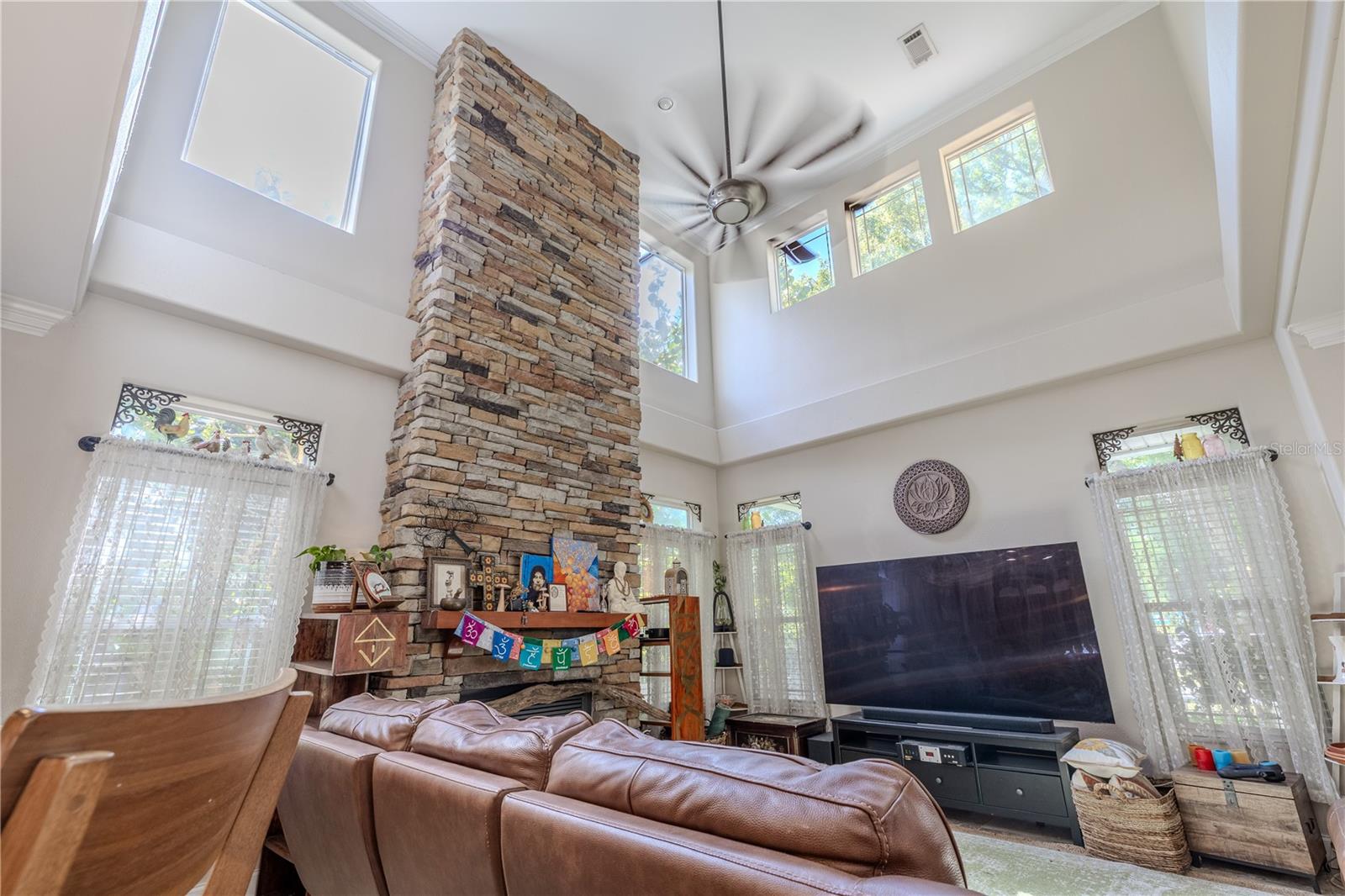 A dramatic two-story living room that blends comfort with architectural flair.Soaring ceilings and clerestory windows flood the space with natural light, while a stunning floor-to-ceiling stacked stone gas fireplace creates a cozy and commanding focal point. Custom transom windows with decorative iron accents and soft sheers add charm and elegance, complementing the room’s open flow to the dining and kitchen areas. With ample space for oversized seating and media, this inviting living area is perfect for everyday living and entertaining alike.