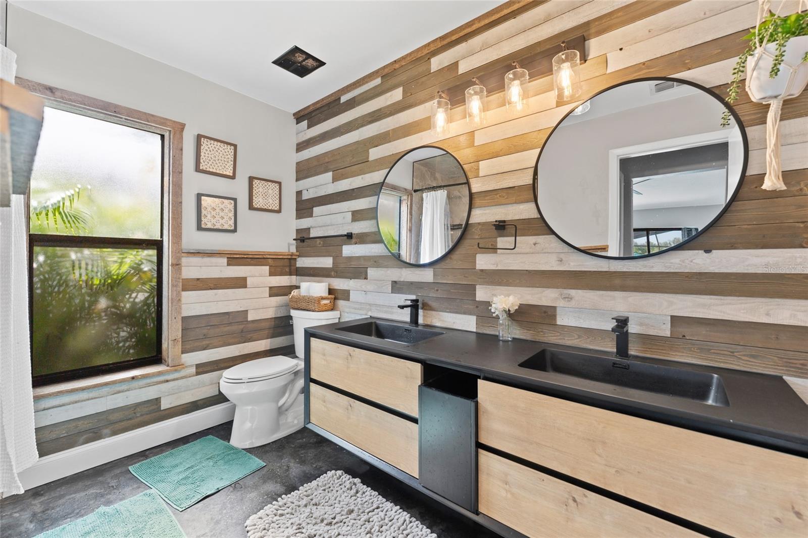 Rustic meets modern in this beautifully renovated bathroom with floating double vanities, black fixtures, round mirrors, and designer lighting. A must-see upgrade!