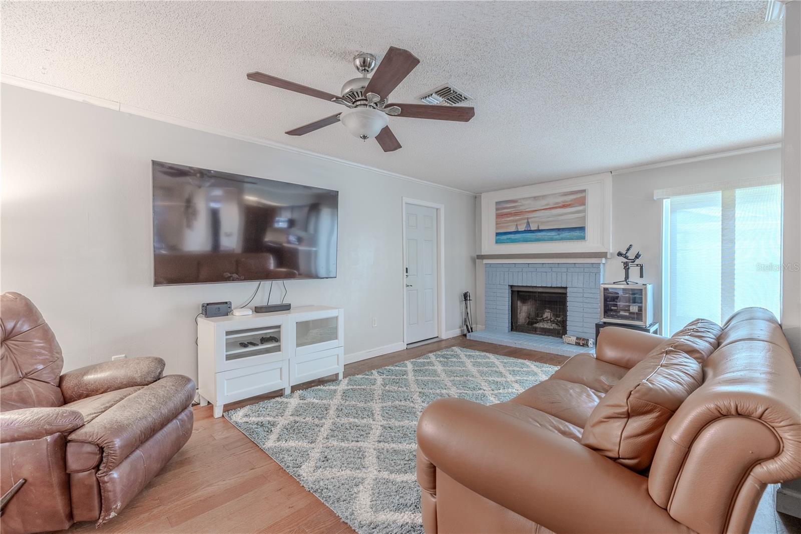 This cozy family room features hardwood floors, a wood-burning fireplace with a painted brick surround and raised hearth—creating a charming focal point for the space. Sliding glass doors allow natural light to fill the room while providing easy access to the outdoor living area. The ceiling fan adds comfort year-round, and the neutral walls and flooring create a warm and versatile backdrop. With ample wall space for a large TV and a seamless flow to the rest of the home, this is a perfect spot for relaxing or entertaining.