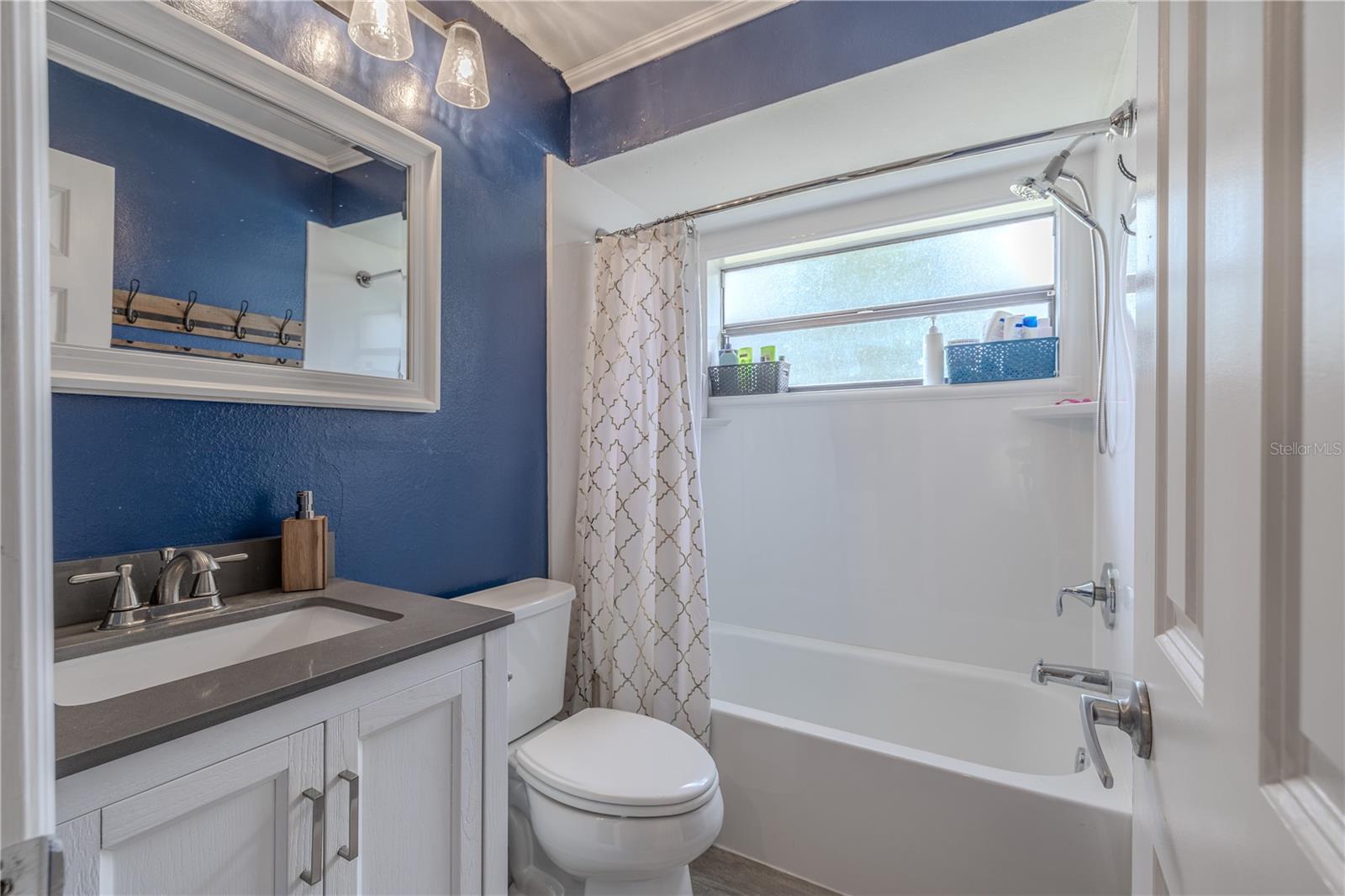 Full bathroom 2 features a clean, modern design with a tub/shower combination, perfect for guests or family use. A wide horizontal window allows natural light to brighten the space while maintaining privacy. The white vanity includes shaker-style cabinetry, a sleek solid-surface countertop, and brushed nickel fixtures. Bold accent walls add a touch of personality, while updated lighting and hardware provide a polished finish.