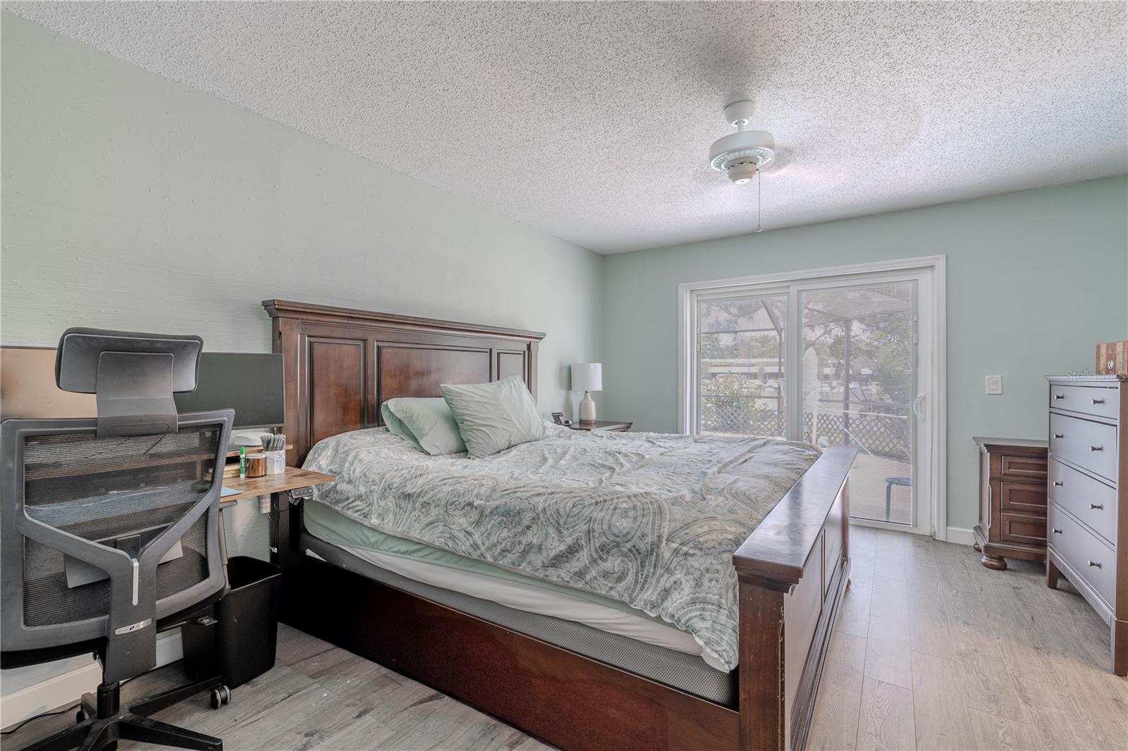 This spacious primary bedroom features a soft color palette, wood-look laminate flooring, and plenty of natural light streaming in through large sliding glass doors that lead to the lanai. A ceiling fan overhead adds comfort and circulation.The room includes a generous walk-in closet and an en-suite bath for added convenience and privacy. With ample floor space and a versatile layout, this bedroom offers comfort, functionality, and flexibility to suit your lifestyle.