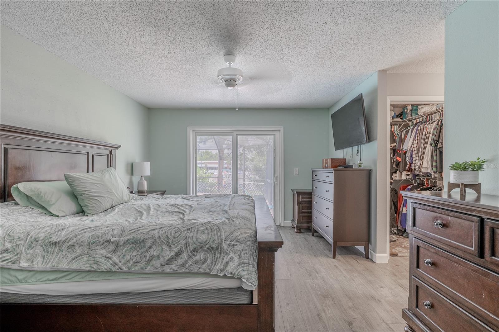 This spacious primary bedroom features a soft color palette, wood-look laminate flooring, and plenty of natural light streaming in through large sliding glass doors that lead to the lanai. A ceiling fan overhead adds comfort and circulation.The room includes a generous walk-in closet and an en-suite bath for added convenience and privacy. With ample floor space and a versatile layout, this bedroom offers comfort, functionality, and flexibility to suit your lifestyle.