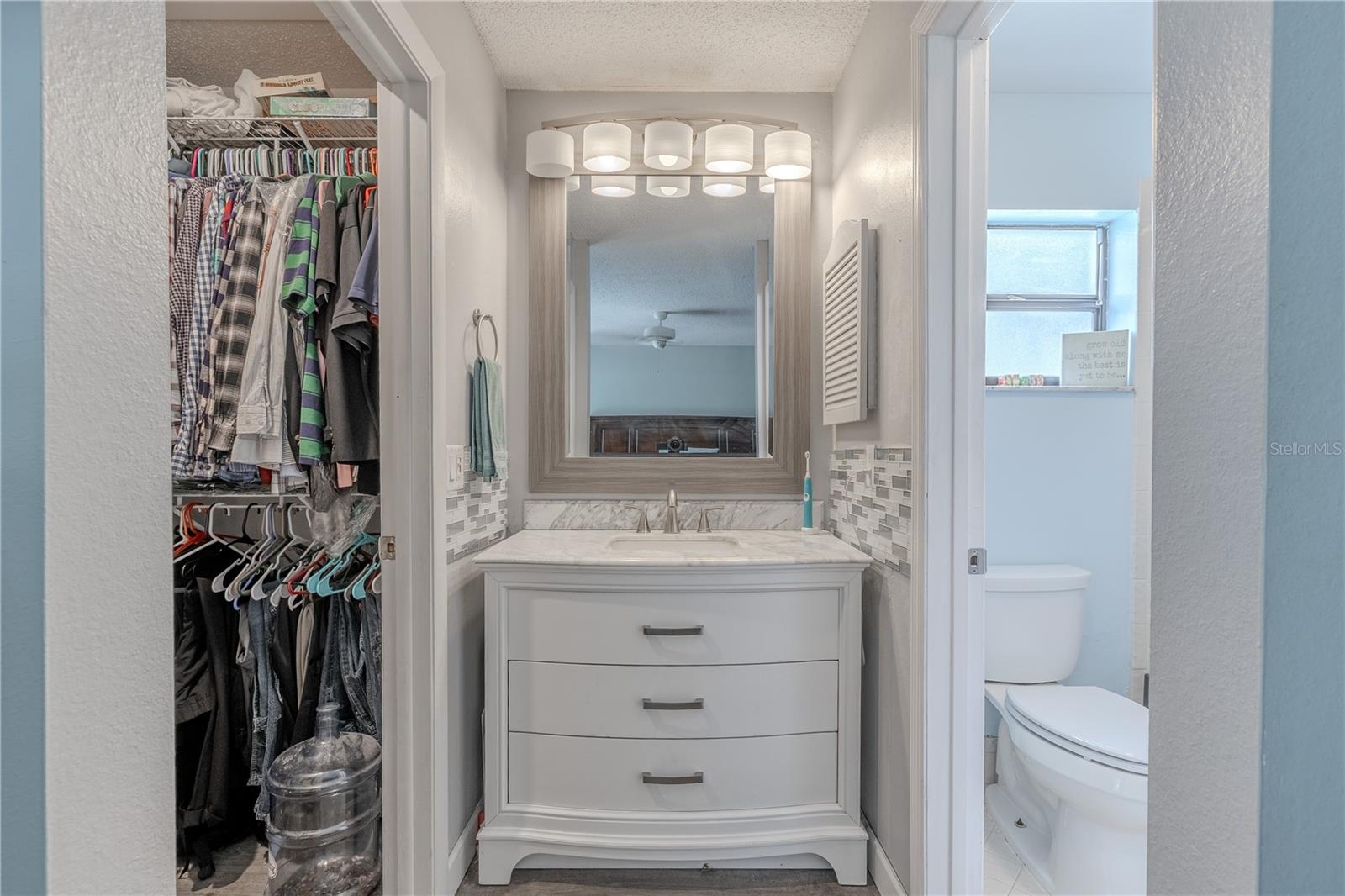 The primary bathroom vanity features a sophisticated curved-front design with crisp white cabinetry,  warm brushed nickel hardware, and a framed mirror with modern lighting above. The vanity is topped with beautiful stone countertops, decorative tile accents that add visual interest to the backsplash creating a luxurious, cohesive look. The bathroom includes a walk-in shower with sliding glass doors and a tiled surround,  Bright and inviting, this space offers a clean, well-maintained feel ideal for everyday comfort.