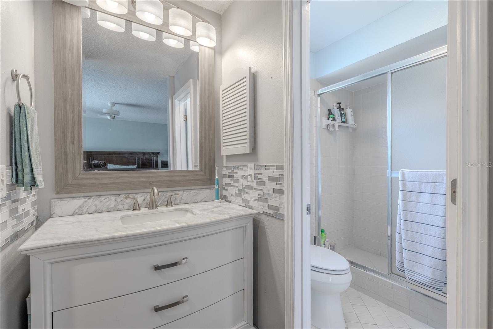 The primary bathroom vanity features a sophisticated curved-front design with crisp white cabinetry,  warm brushed nickel hardware, and a framed mirror with modern lighting above. The vanity is topped with beautiful stone countertops, decorative tile accents that add visual interest to the backsplash creating a luxurious, cohesive look. The bathroom includes a walk-in shower with sliding glass doors and a tiled surround,  Bright and inviting, this space offers a clean, well-maintained feel ideal for everyday comfort.