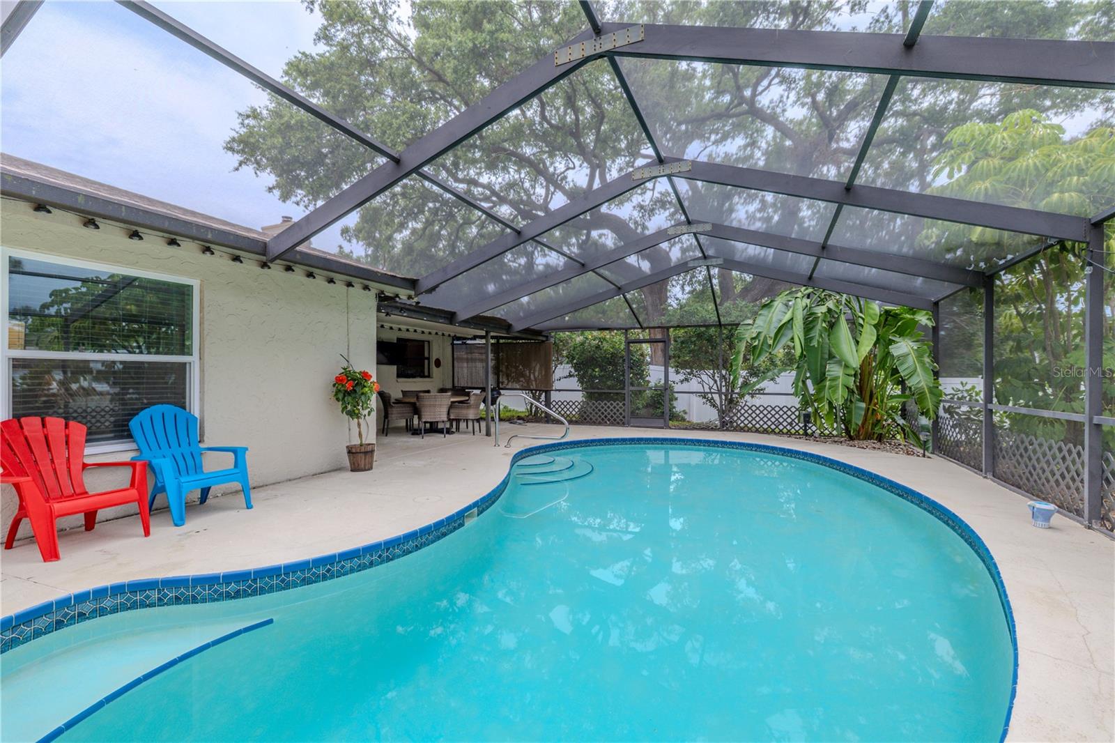 Enjoy year-round outdoor living with this sparkling screened-in pool, surrounded by a spacious lanai and lush tropical landscaping. The curved pool design features built-in steps for easy entry and a tile-accented border that adds a polished touch. The expansive covered area offers plenty of room for dining and entertaining, while the screened enclosure keeps the space shaded, breezy, and bug-free. Whether you’re hosting guests or relaxing poolside, this private outdoor retreat is the perfect place to unwind.