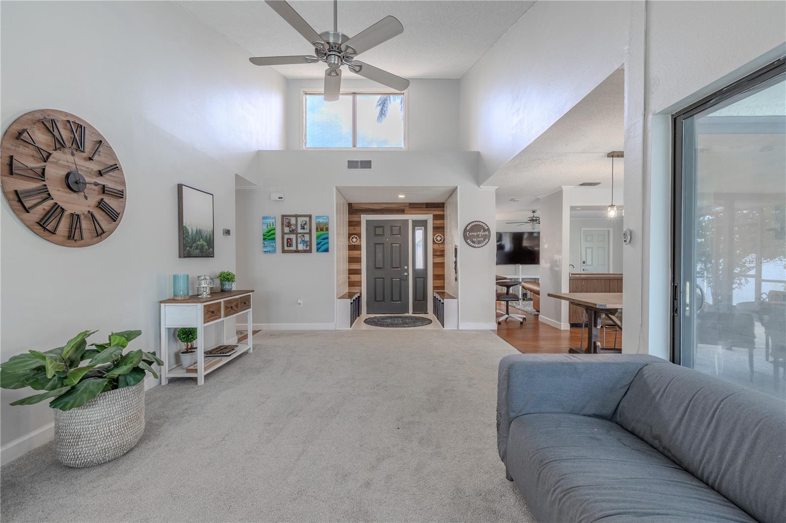 This spacious living room features dramatic vaulted ceilings creates an impressive sense of height and openness. The room is enhanced by abundant natural light streaming through a large upper window, while a ceiling fan provides comfortable air circulation.The neutral carpet flooring flows seamlessly throughout the space, connecting this living area to the adjacent kitchen and dining spaces in a true open-concept design. The layout offers excellent sightlines across the main living areas, creating a cohesive and welcoming environment.