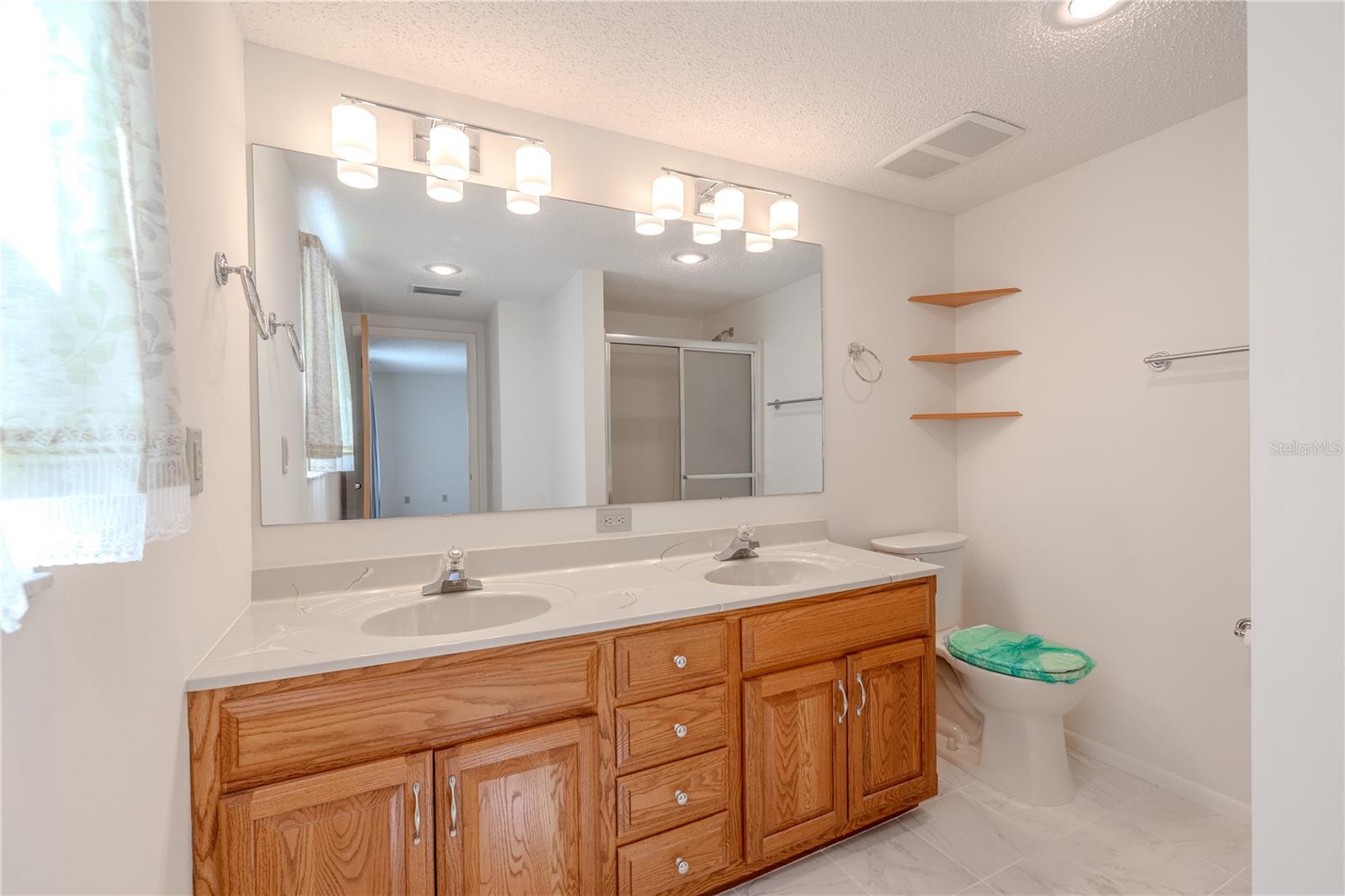 The primary ensuite bathroom offers both space and functionality, featuring a large dual-sink vanity with abundant storage and a wide mirror framed by modern lighting. Neutral tile flooring and soft white walls provide a clean, timeless backdrop. A step-in shower with sliding glass doors adds everyday convenience, while built-in corner shelving offers extra space for toiletries or decorative touches. Located just off the spacious primary bedroom, this ensuite delivers a practical and private retreat.