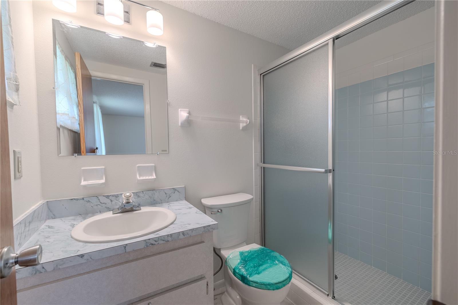 Bathroom 2 offers a bright and functional space with classic finishes and a practical layout. It features a walk-in shower with frosted glass sliding doors and soft blue tile surround, adding a subtle pop of color. The single-sink vanity includes a marble-look countertop with integrated backsplash and ample under-sink storage. A large mirror with modern lighting completes the space, making it ideal for guests, as an en suite for Bedroom 2. Clean, neutral tones throughout offer a timeless backdrop ready for your personal touch.