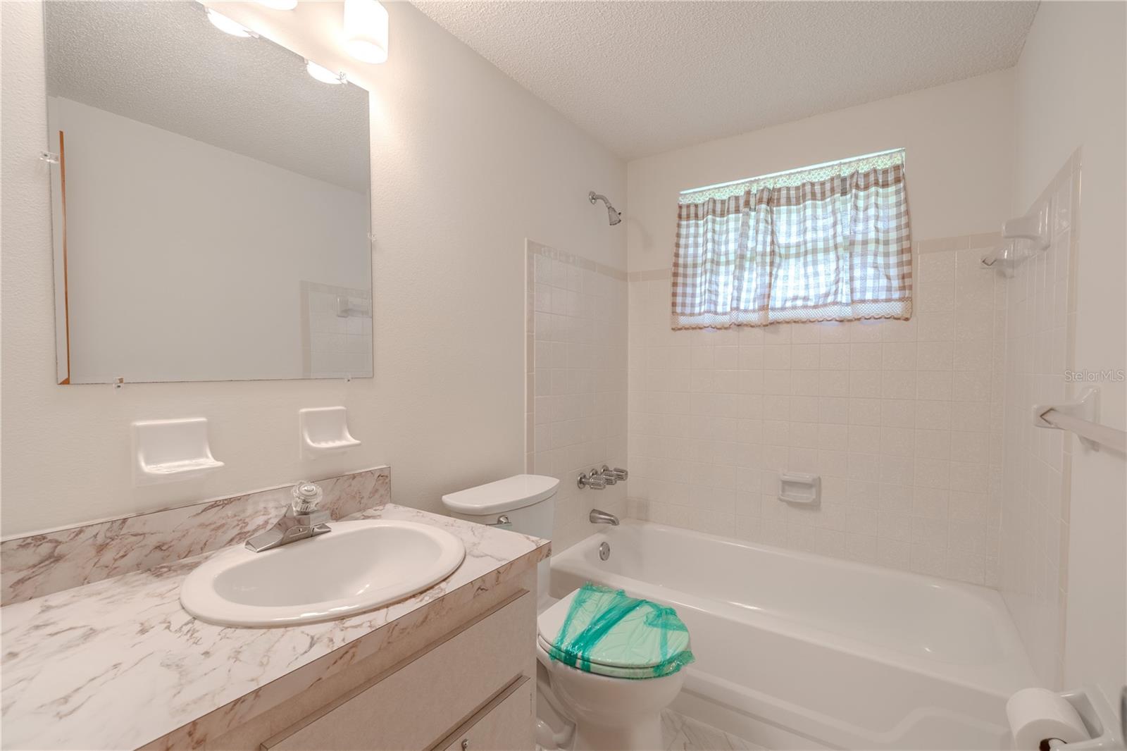 Bathroom 3 features a classic and functional design, complete with a tub-shower combo and full tile surround—ideal for guests or everyday use. The single-sink vanity offers ample counter space with a marble-look finish and under-sink storage. A large wall-mounted mirror and modern lighting add brightness to the space, while a small window above the tub allows natural light to filter in. With its neutral tones and practical layout, this bathroom is both charming and convenient.