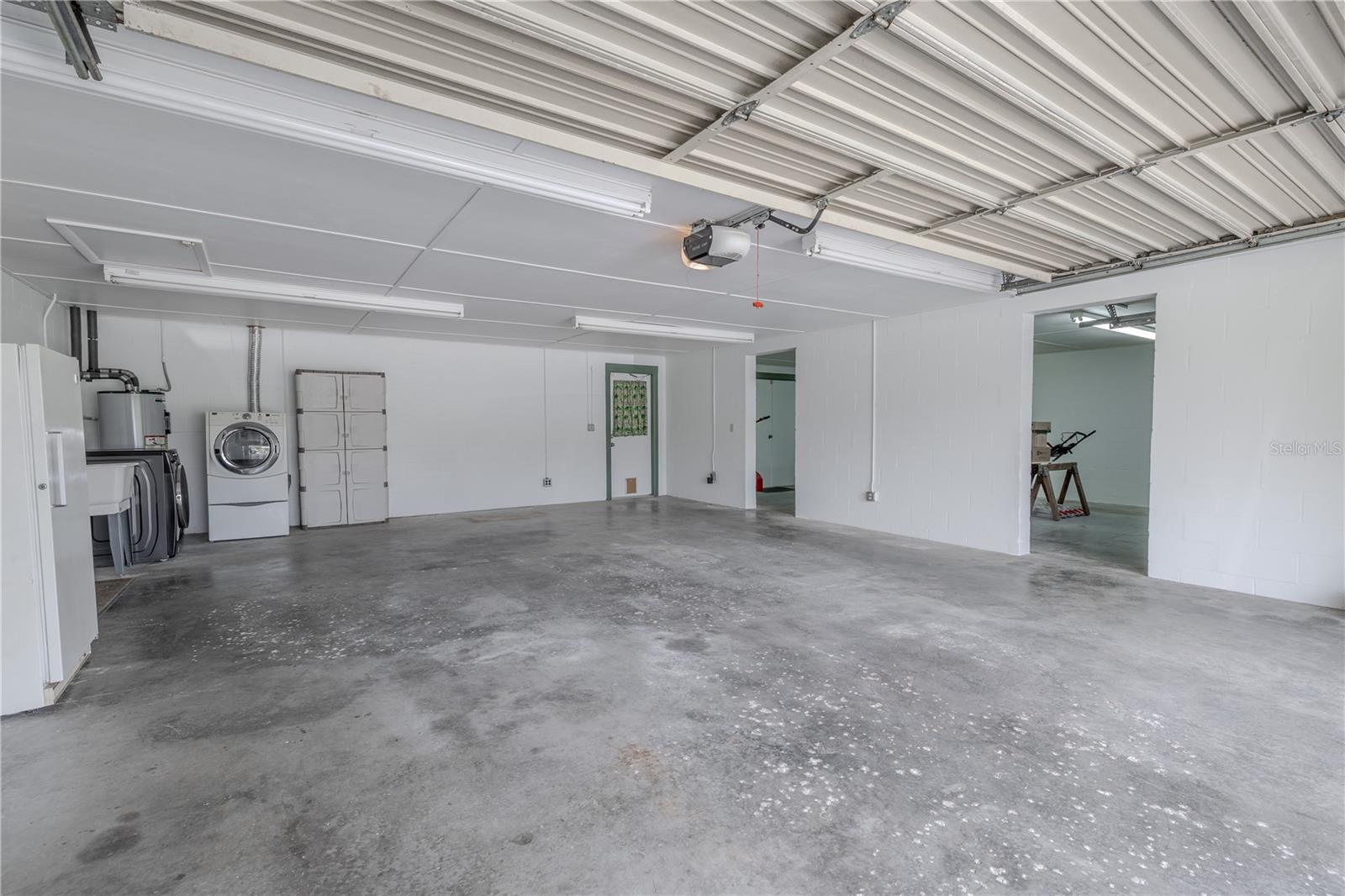 This oversized two-car garage offers ample space for vehicles, storage, or a workshop setup in a bright, clean environment, enhanced by multiple overhead fluorescent light fixtures for excellent visibility. The space includes a dedicated laundry area with washer, dryer, utility sink, and water heater, all neatly tucked along the back wall. There's also direct access to the interior of the home and backyard, along with a convenient side storage or workroom. A solid choice for anyone needing both functionality and extra elbow room.