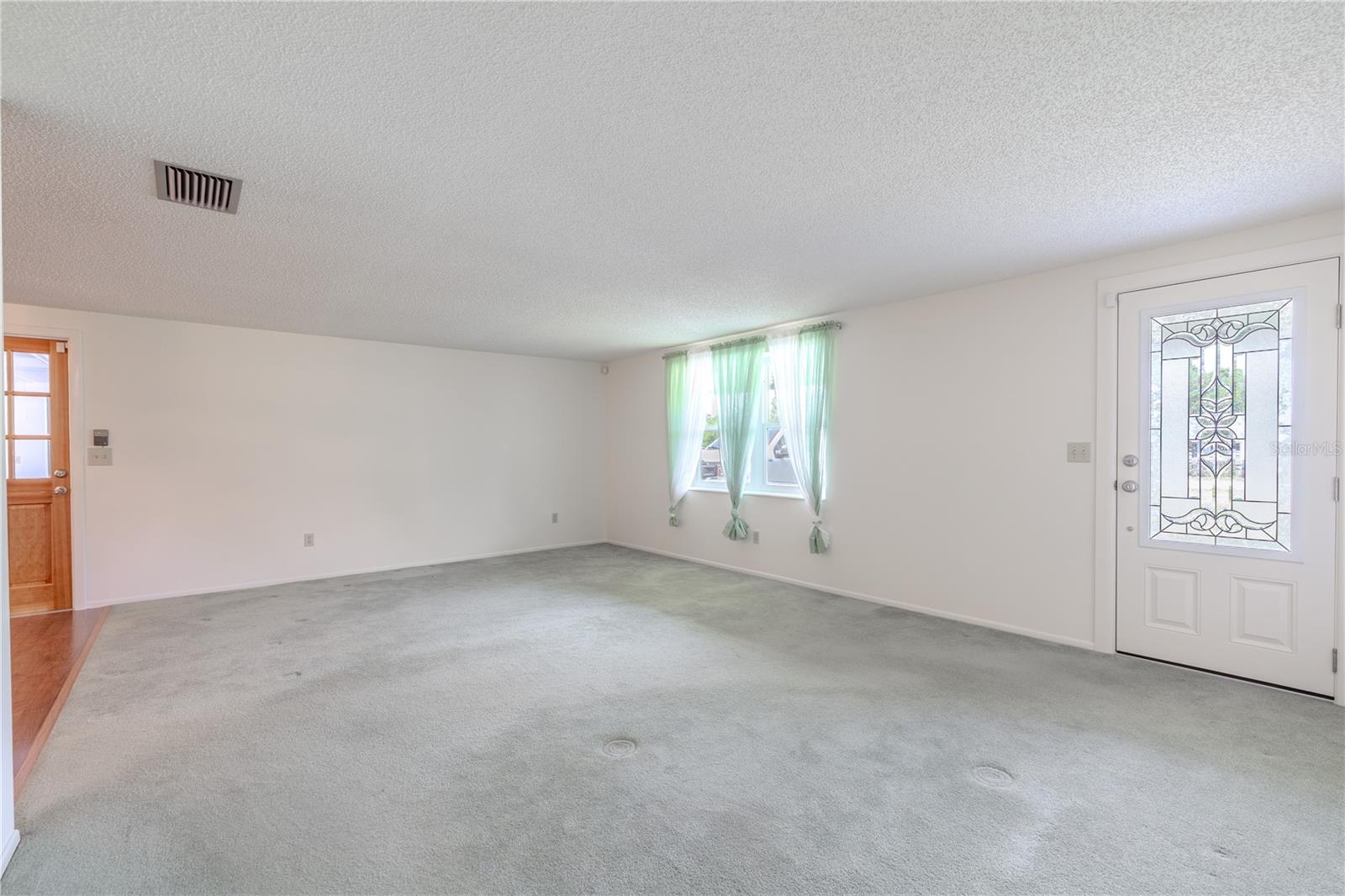 This spacious family room offers a clean and versatile canvas, ready to accommodate a variety of furniture layouts and design styles. A large front-facing window fills the room with natural light, while the neutral wall color and soft carpet flooring enhance the sense of openness. The decorative glass-panel front door adds architectural interest and additional brightness to the space.