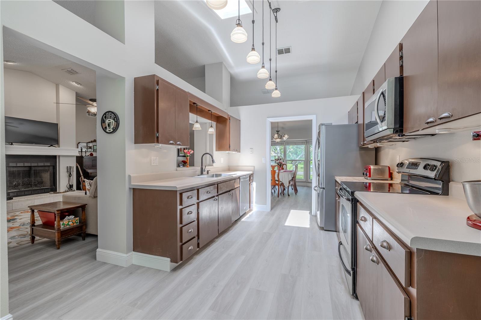 The kitchen is both bright and functional, offering a spacious galley-style layout with excellent flow and visibility to the main living and dining areas. Vaulted ceilings with dual skylights and a row of pendant lights create a light-filled, airy space ideal for cooking and entertaining.