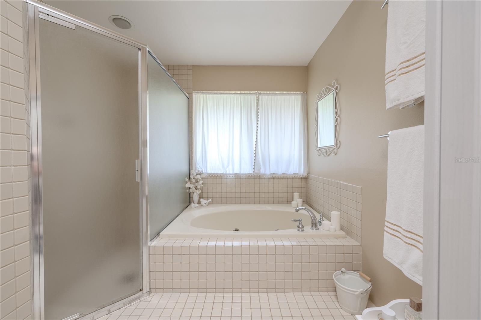 Primary bathroom: The step-up soaking tub,  beneath a wide window with soft natural light, invites you to unwind at the end of the day. A separate walk-in shower with frosted glass enclosure offers added convenience, while neutral tile flooring and soft beige tones create a clean and calming spa-like atmosphere.