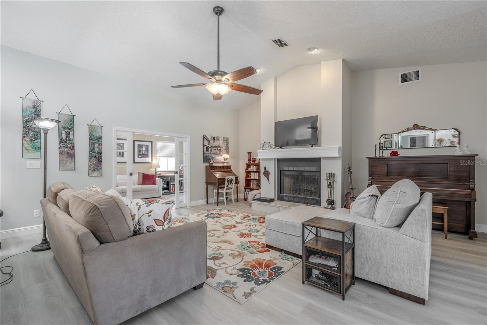 This inviting living room boasts vaulted ceilings and recessed lighting that create a spacious and airy feel. A classic wood-burning fireplace with a raised hearth and elegant mantle anchors the room, offering warmth and character. Freshly painted walls in a soft, neutral tone complement the luxury vinyl plank flooring installed in 2025, giving the space a clean and updated loo