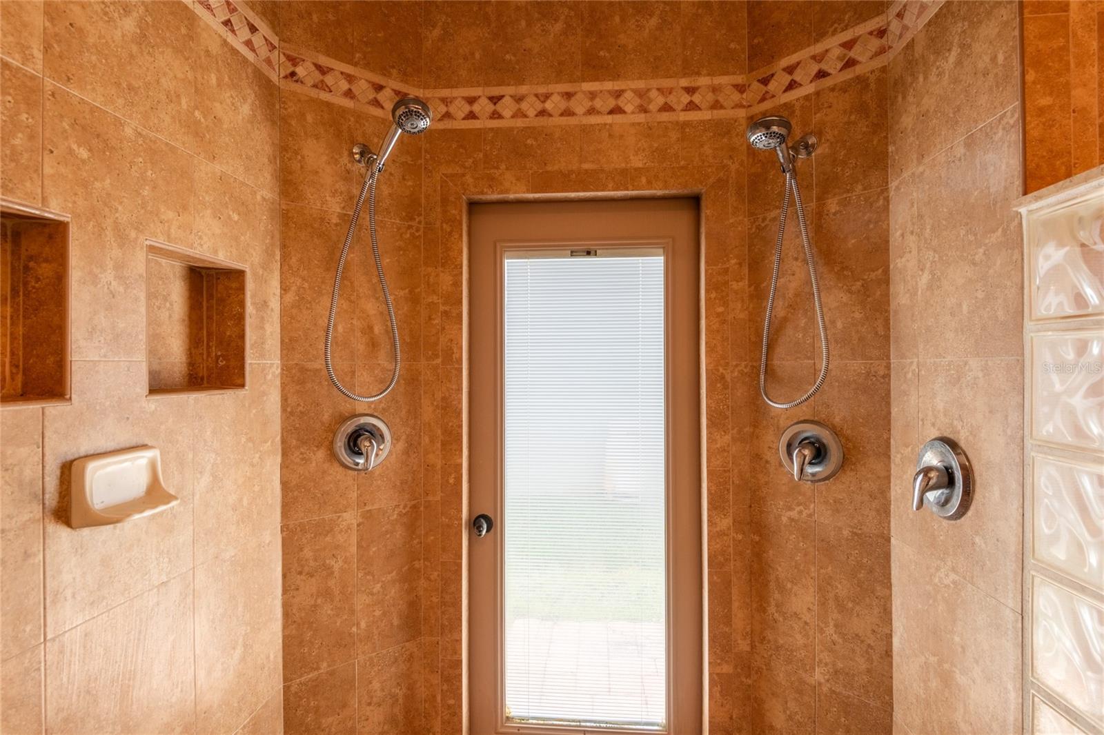 Master shower with dual shower heads and dustless window blind door leading to outside.