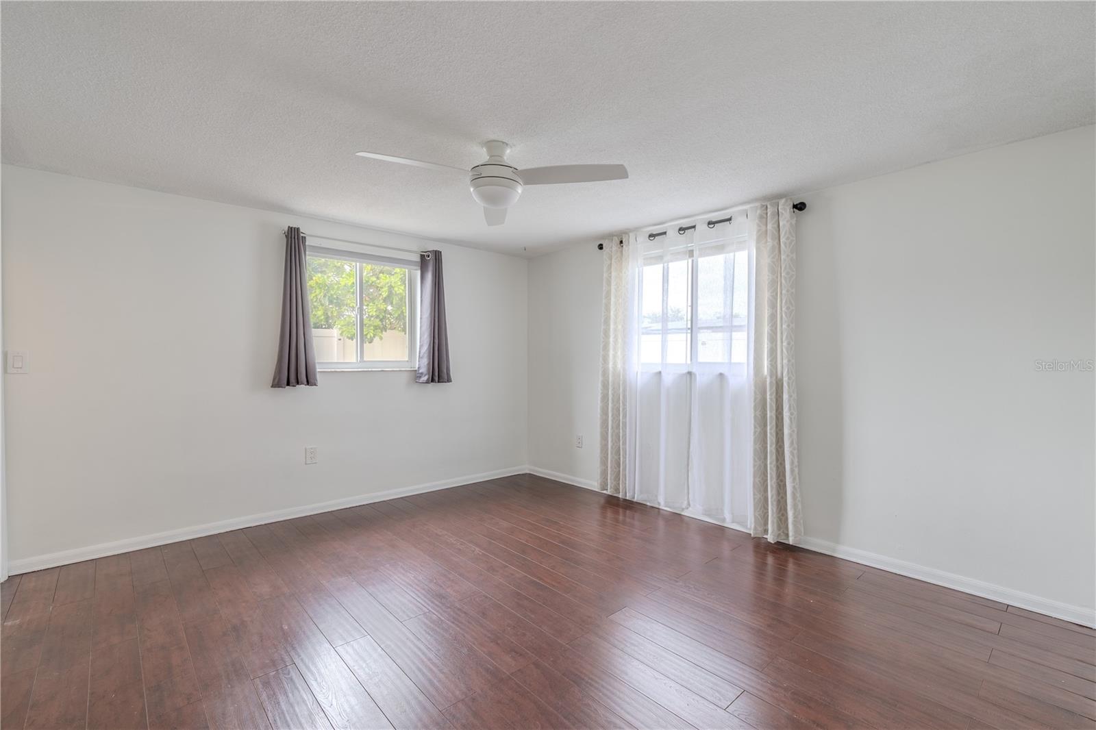 The spacious primary suite offers a quiet retreat with rich wood-look flooring, soft neutral walls, and an abundance of natural light filtering in through dual windows. A modern ceiling fan ensures year-round comfort, while the layout provides plenty of room for a king-sized bed and additional furniture.