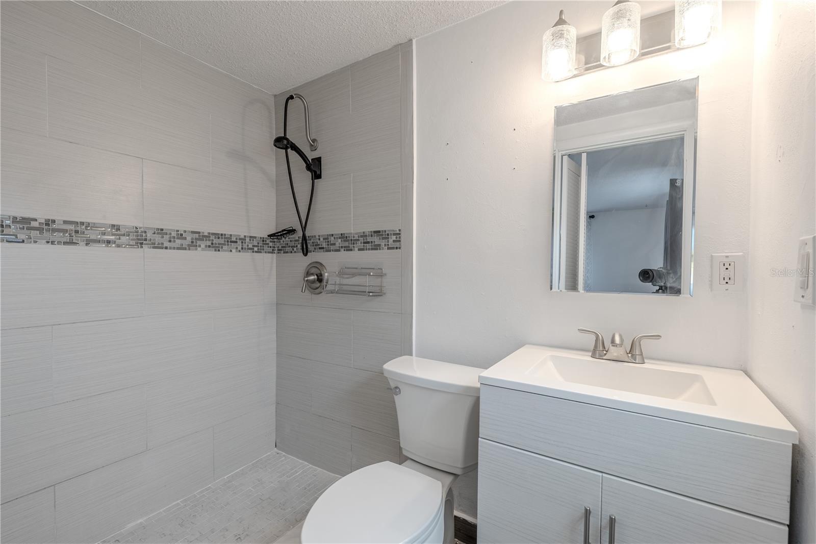 The private en suite bath has been stylishly updated with a sleek walk-in shower featuring oversized tile, mosaic accent banding, and a handheld fixture for added convenience. The contemporary vanity offers clean lines and ample storage, topped with a seamless integrated sink and framed by a mirror and trio of glass lights.