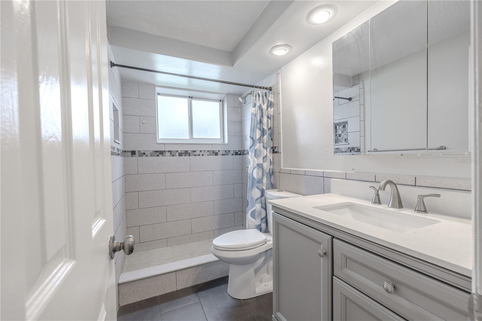 A sleek vanity with a solid-surface countertop provides generous counter space and storage, while the wide mirror and overhead lighting keep the room bright and practical. The soft, neutral finishes and contemporary fixtures create a spa-like feel that complements the home’s cohesive design.