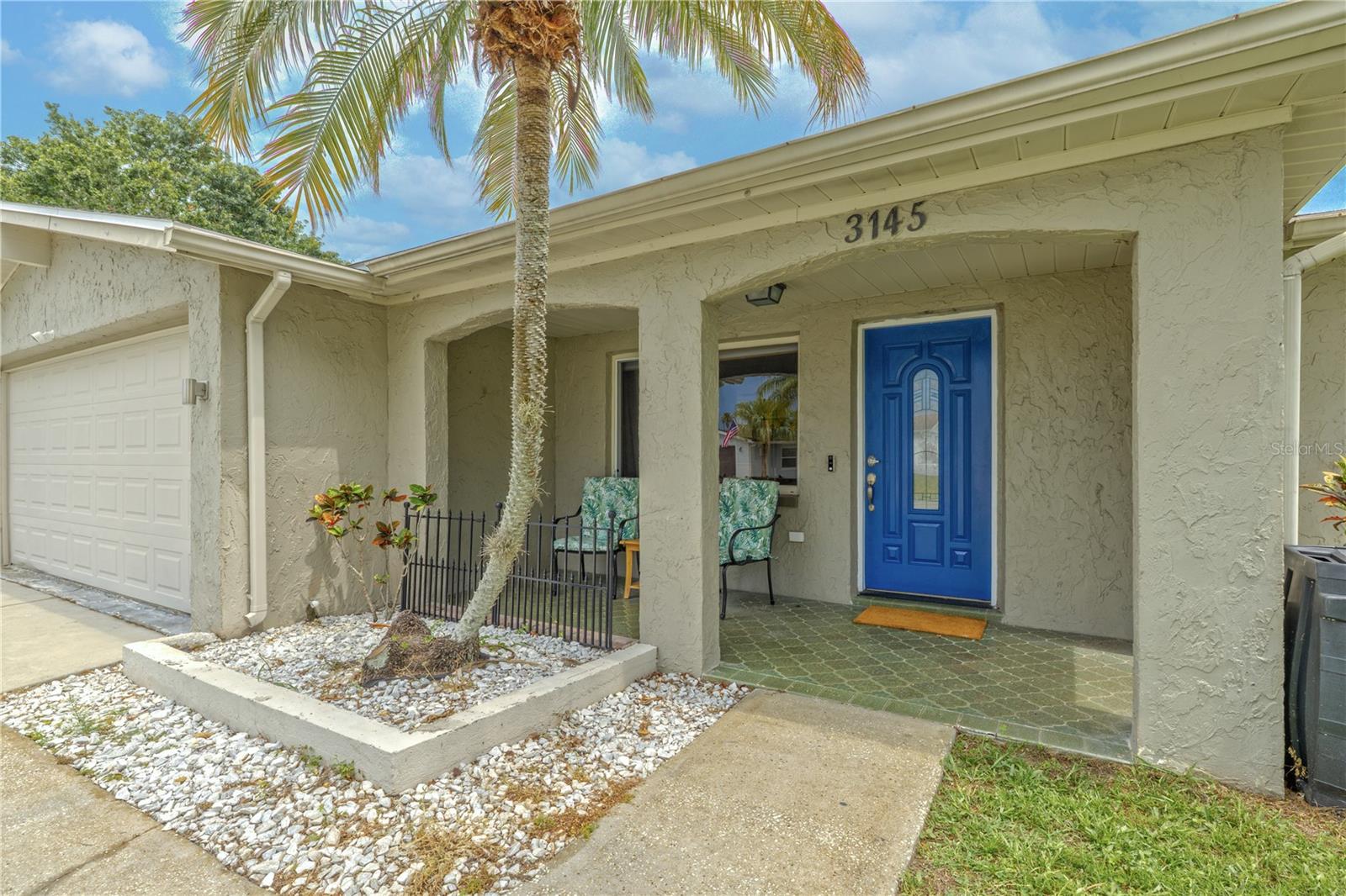 A bold, eye-catching cobalt blue front door sets the tone on this front porch, welcoming guests with personality and style. The covered entryway offers a shady retreat with space for seating—perfect for enjoying your morning coffee or chatting with neighbors in the fresh air.Textured stucco arches frame the porch area, while a decorative tile-style concrete floor adds character underfoot. A cozy seating nook with room for a small table and chairs overlooks a neatly landscaped garden bed lined with white stone and tropical palms, creating a serene first impression. This is a front porch that says, “Come on in and stay awhile.”