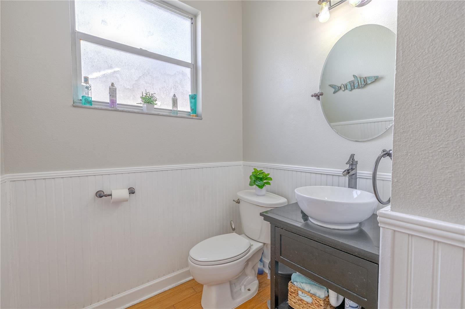 The first-floor half bath offers a fresh and stylish design, featuring classic white beadboard wainscoting and a soft gray upper wall that adds dimension and contrast. A round mirror is set above a contemporary vessel sink, mounted on a sleek black vanity with open shelving for added functionality. A frosted glass window allows in natural light while maintaining privacy.