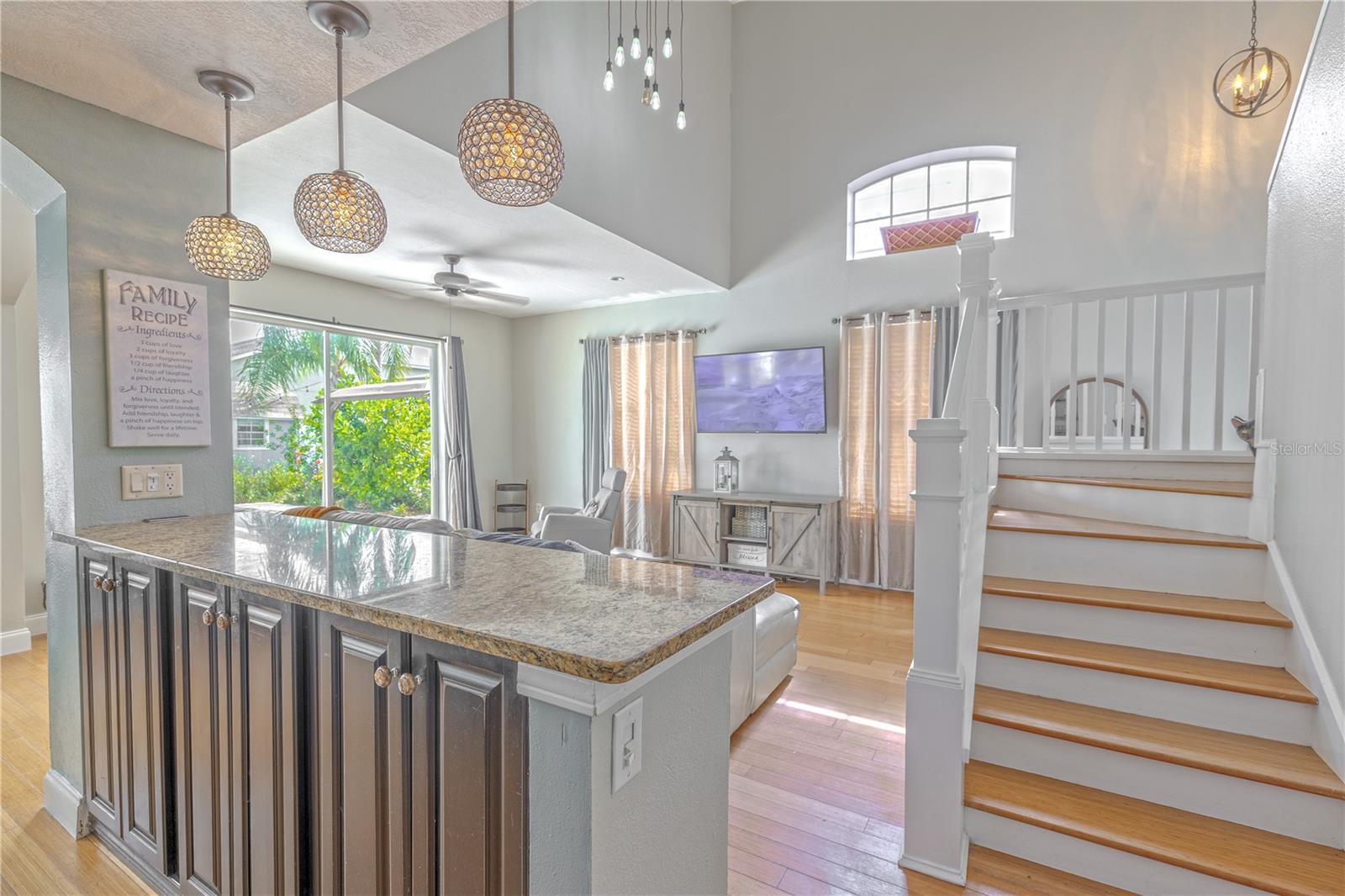From the kitchen, the view opens to a bright and spacious living area, framed by soaring ceilings and a flood of natural light from tall windows and a large sliding glass door that leads to the backyard oasis. A granite-topped bar provides a perfect visual and functional transition between the kitchen and living room, with pendant lighting above adding a modern design touch.To the right, a crisp white staircase with wood treads and a sleek balustrade leads to the second floor, accented by a high arched window that adds both architectural interest and additional light. Overhead, a multi-pendant chandelier adds a stylish statement, enhancing the home's airy and open feel.