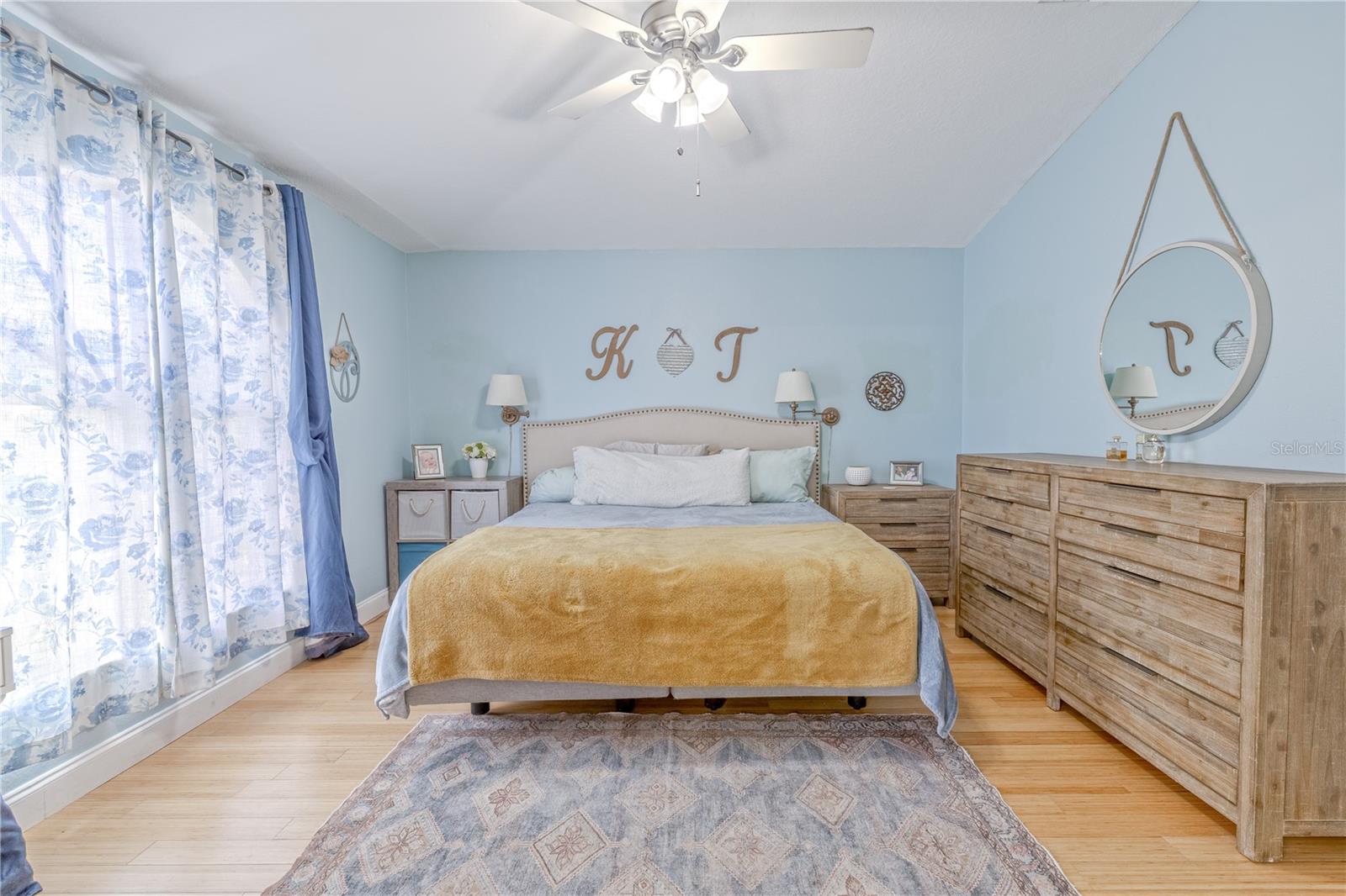 The spacious primary bedroom(with ensuite bath)  has soft natural light, vaulted ceilings, and calming hues that enhance the sense of openness. Bamboo flooring adds warmth and continuity, while a ceiling fan overhead ensures year-round comfort. The layout provides ample space for a king-sized bed and accompanying furniture, with thoughtful wall placement ideal for both aesthetics and function. A large window dressed in light-filtering drapes brightens the room with gentle daylight while maintaining privacy.With its clean lines, generous proportions, and tranquil atmosphere, this primary suite is the perfect place to unwind at the end of the day.