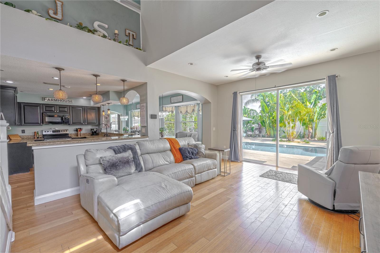 The sun-soaked living room perfectly balances warmth, comfort, and style. With soaring ceilings and wide-open sightlines to the kitchen and pool area, this central gathering space invites both relaxed lounging and vibrant entertaining. Natural light pours through oversized sliding glass doors, drawing your eyes straight to the sparkling pool and lush tropical landscaping beyond.