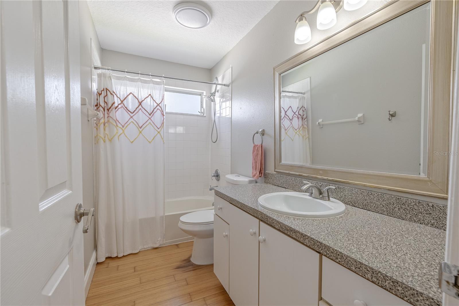 The second full bathroom offers a bright and functional design, featuring a long solid-surface vanity with an under-mount sink and ample counter space. A large framed mirror sits beneath a triple-light fixture, providing even illumination across the room.A combination tub and shower is finished in classic white tile and includes a frosted glass window that brings in natural light while maintaining privacy. Bamboo flooring adds warmth and continuity from the adjacent rooms, while neutral wall tones make this space feel fresh and versatile. Ideal for guests or family use, this bathroom is both practical and inviting.