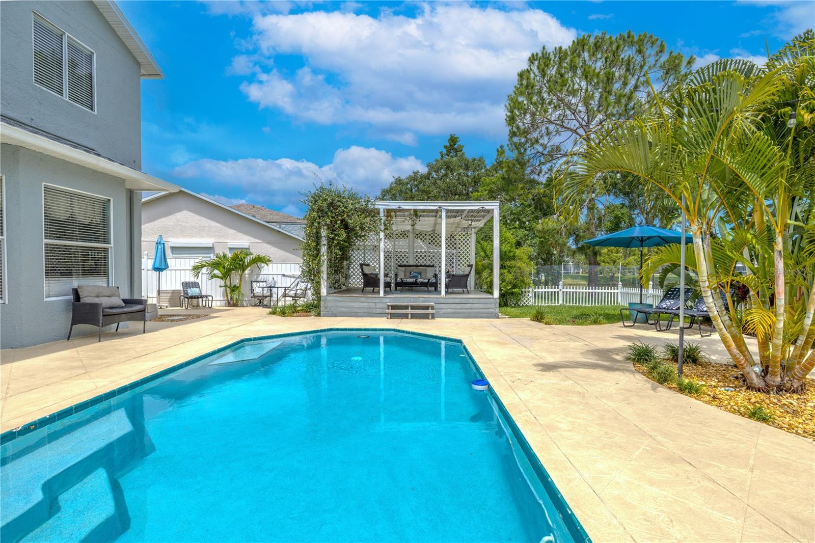Just beyond the pool, a raised pergola with latticework and shaded seating creates an intimate setting for lounging or entertaining.Tropical landscaping adds privacy and a lush, resort-like feel, while multiple seating zones—including shaded lounge chairs and a small patio by the house—invite relaxation under the Florida sun. The expansive pool deck provides ample room for gatherings, grilling, or quiet mornings with a coffee in hand.