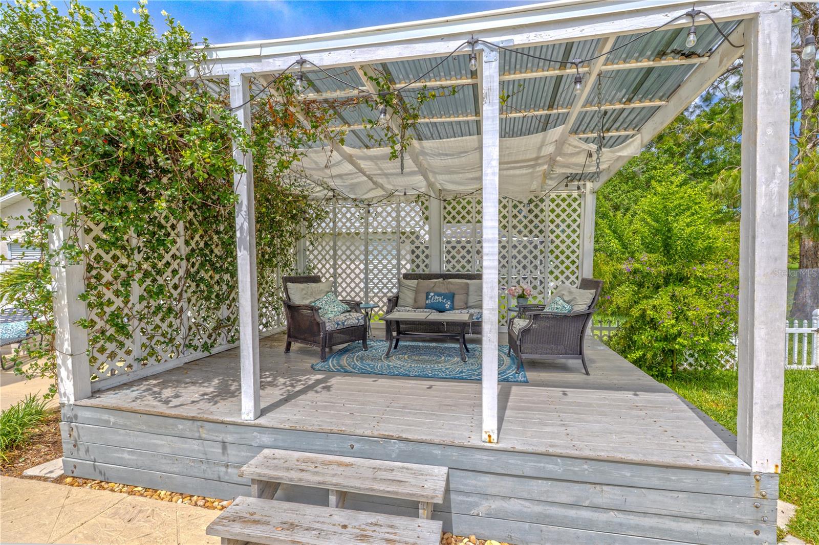 Tucked into the corner of the backyard, this elevated pergola offers a charming escape perfect for both relaxation and entertaining. Climbing vines wrap gracefully around the lattice panels, adding natural privacy and a lush, garden-like atmosphere. The partially covered roof allows soft filtered light to pass through, while string lights overhead create a cozy, ambient glow as the sun sets.Built with wide wooden decking and a few easy steps for access, this versatile space is ideal for lounging with a book, sipping evening cocktails, or hosting guests beside the pool. It’s a picture-perfect retreat that feels like an extension of the home’s indoor living area—only with a breeze and a view.
