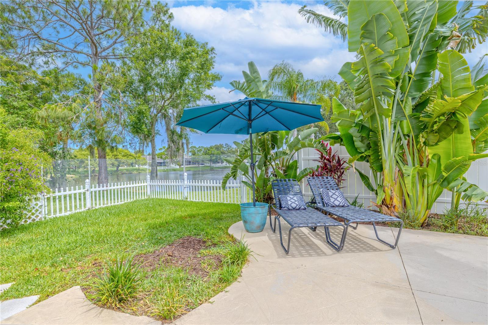 Tucked in a sun-drenched corner of the backyard, this serene spot is perfect for soaking in the Florida sunshine or unwinding with a good book. Ideal placement for chaise lounges to rest on the extended pool deck, shaded by an umbrella and surrounded by swaying palms, colorful tropical foliage, and a lush green lawn.Beyond the white picket fence lies a tranquil pond view, offering a picturesque backdrop of mature trees and reflections dancing on the water. Whether you're looking for morning calm or afternoon sun, this private corner feels like your own slice of paradise.