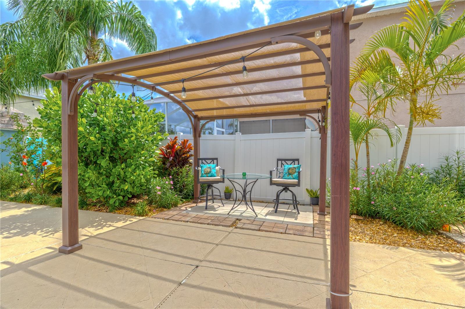 Tucked along the side of the pool deck, this charming pergola adds a cozy retreat framed by lush tropical landscaping. With its curved design and built-in shade canopy, it offers just the right amount of coverage for relaxing outdoors—day or night. String lights are strung overhead, ready to create a soft, inviting ambiance after sunset.Set atop a paved brick pad that defines the space beautifully, this shaded nook is ideal for morning coffee, quiet conversation, or enjoying a cool drink poolside. Surrounded by flowering plants and swaying palms, it’s a peaceful slice of paradise right in your own backyard.