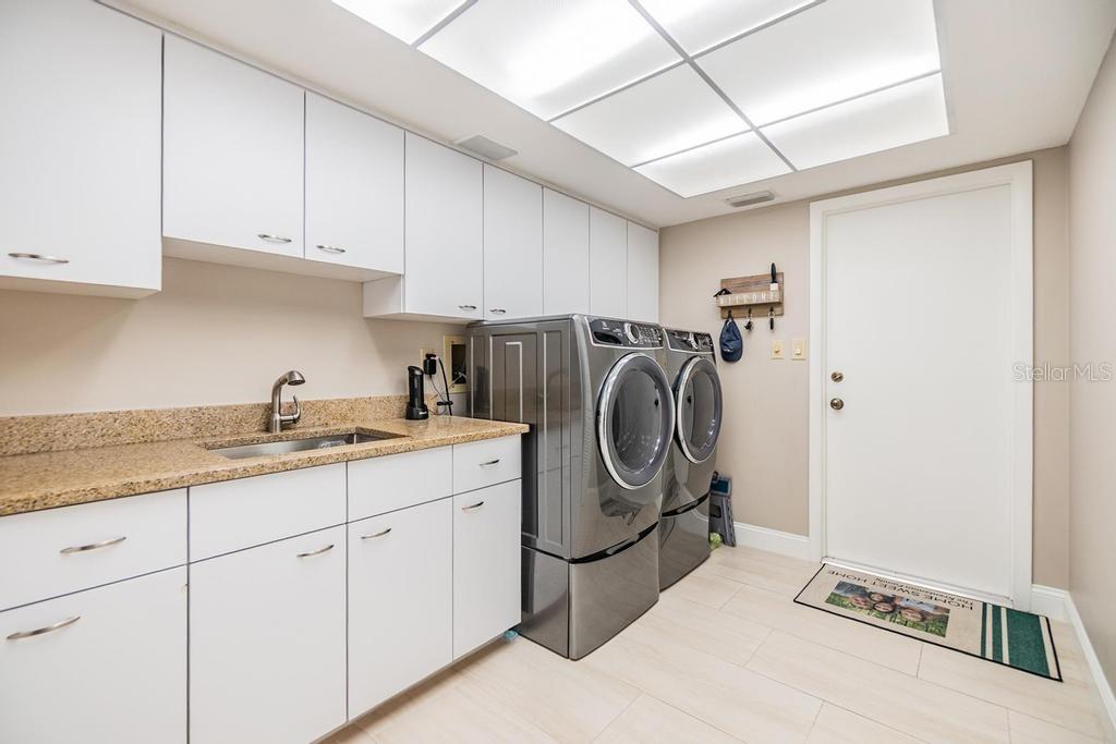 The laundry room is thoughtfully outfitted with a large-capacity washer and dryer on pedestals, a stainless-steel sink, granite folding counter, and generous built-in storage, making everyday chores a breeze