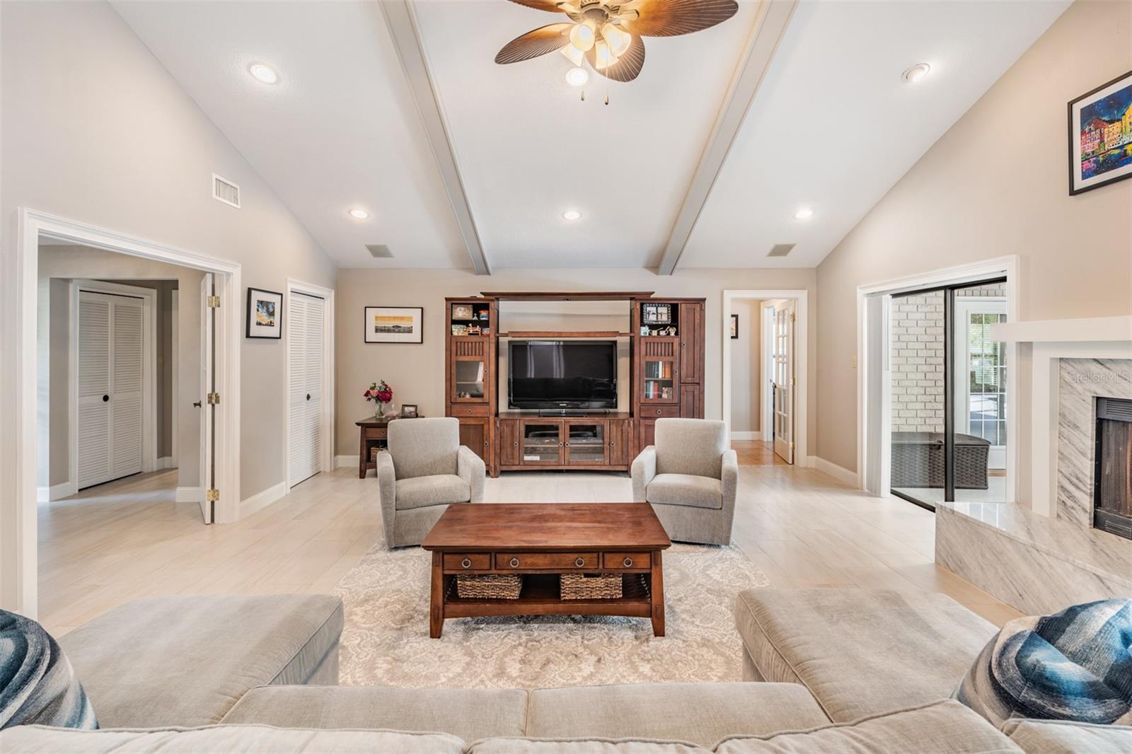 The expansive living room draws you inside with a warm and sophisticated ambiance, accented by neutral tones, gorgeous tile flooring, and a vaulted ceiling with stylish wood beams. A gas fireplace with marble accents serves as a striking focal point, flanked by two sliding glass doors that open to the pool area.