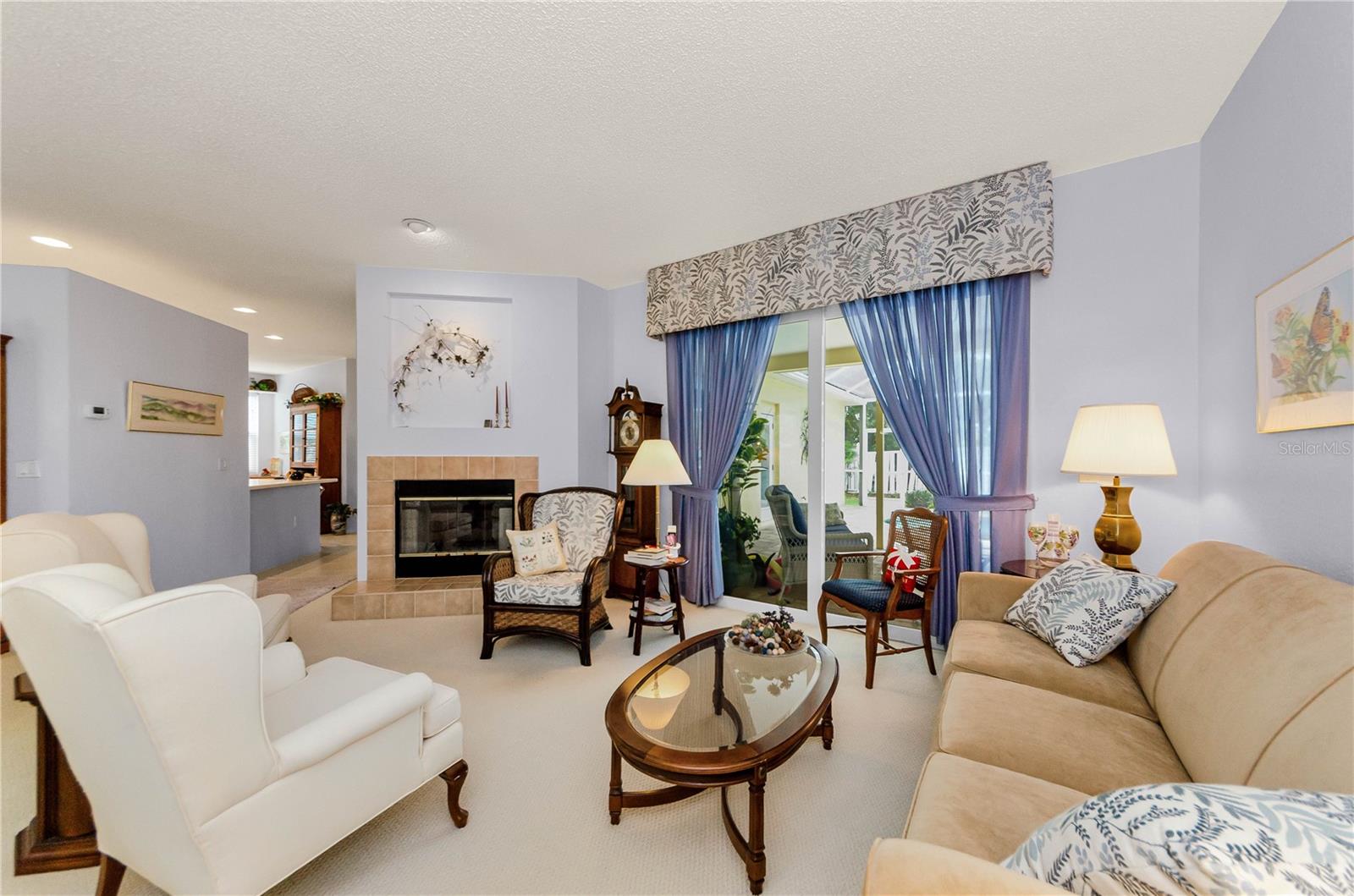 Wide open view of living room with double sided fireplace and view of kitchen to the left, pool through glass sliding doors, and Primary Bedroom to the right.